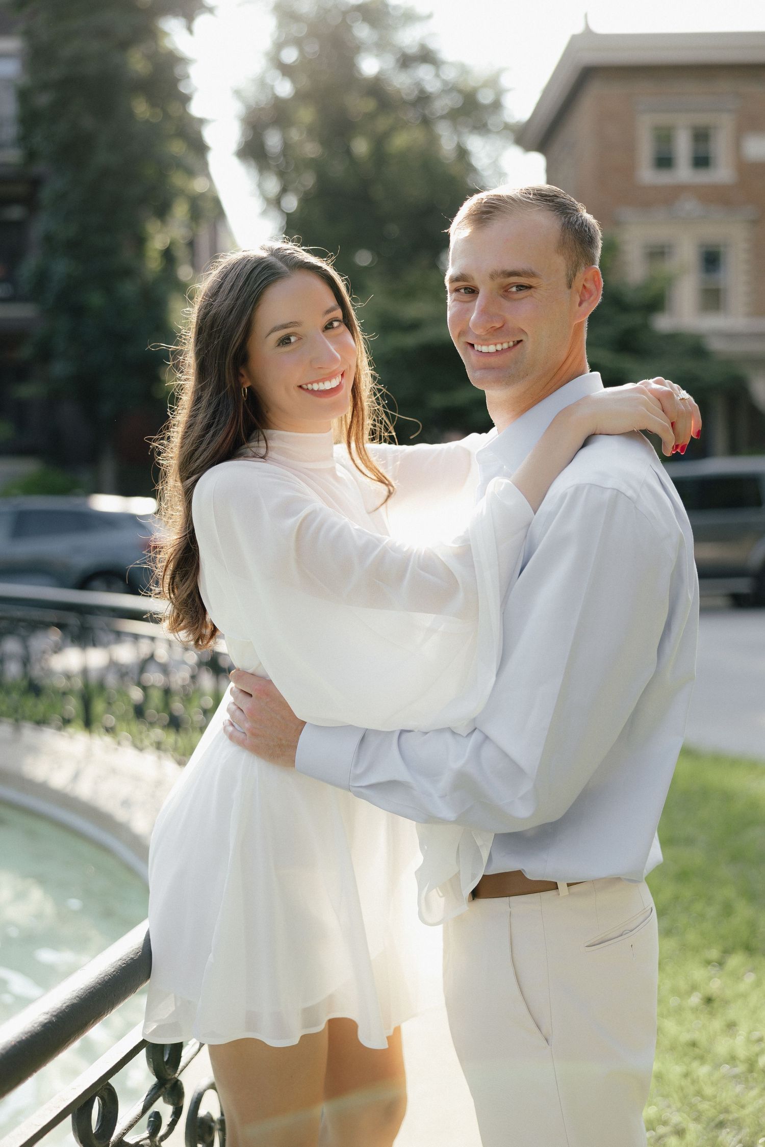 Happy couple embracing outdoors near a fountain, woman in white dress with arms around mans shoulders.