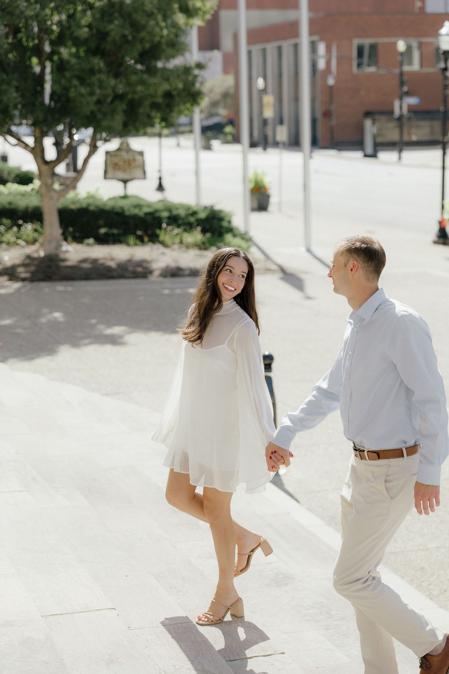 A smiling couple holds hands while walking on a sunny urban sidewalk, woman wearing a white flowy dress.
