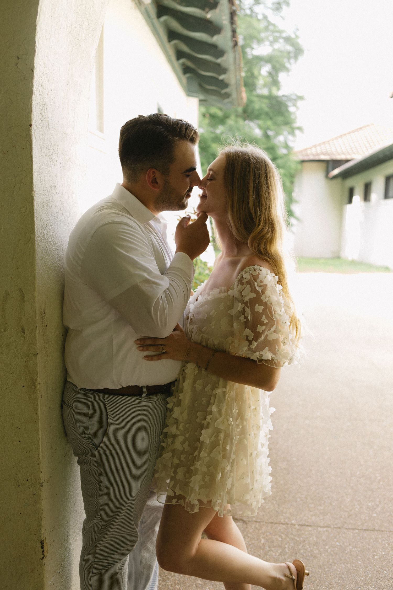Romantic couple in white outfits sharing an intimate moment leaning against a wall outdoors.