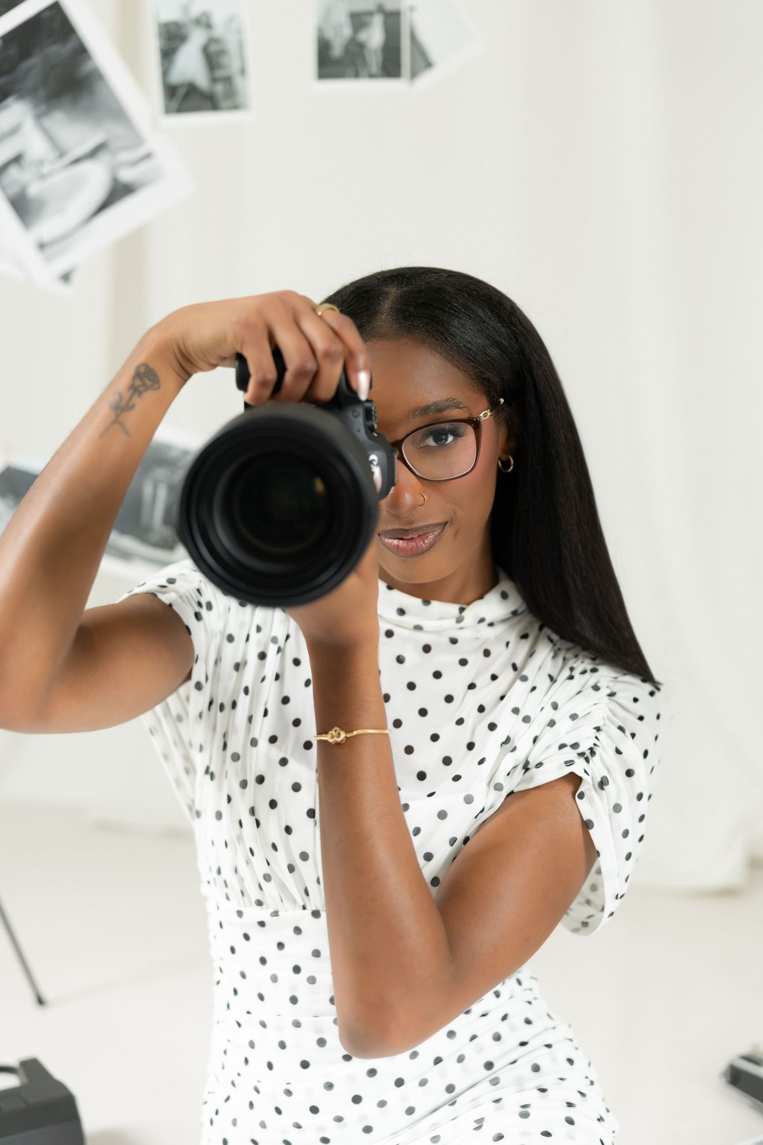 Young Black female photographer in polka dot dress and glasses holding DSLR camera in bright studio with photos hanging.