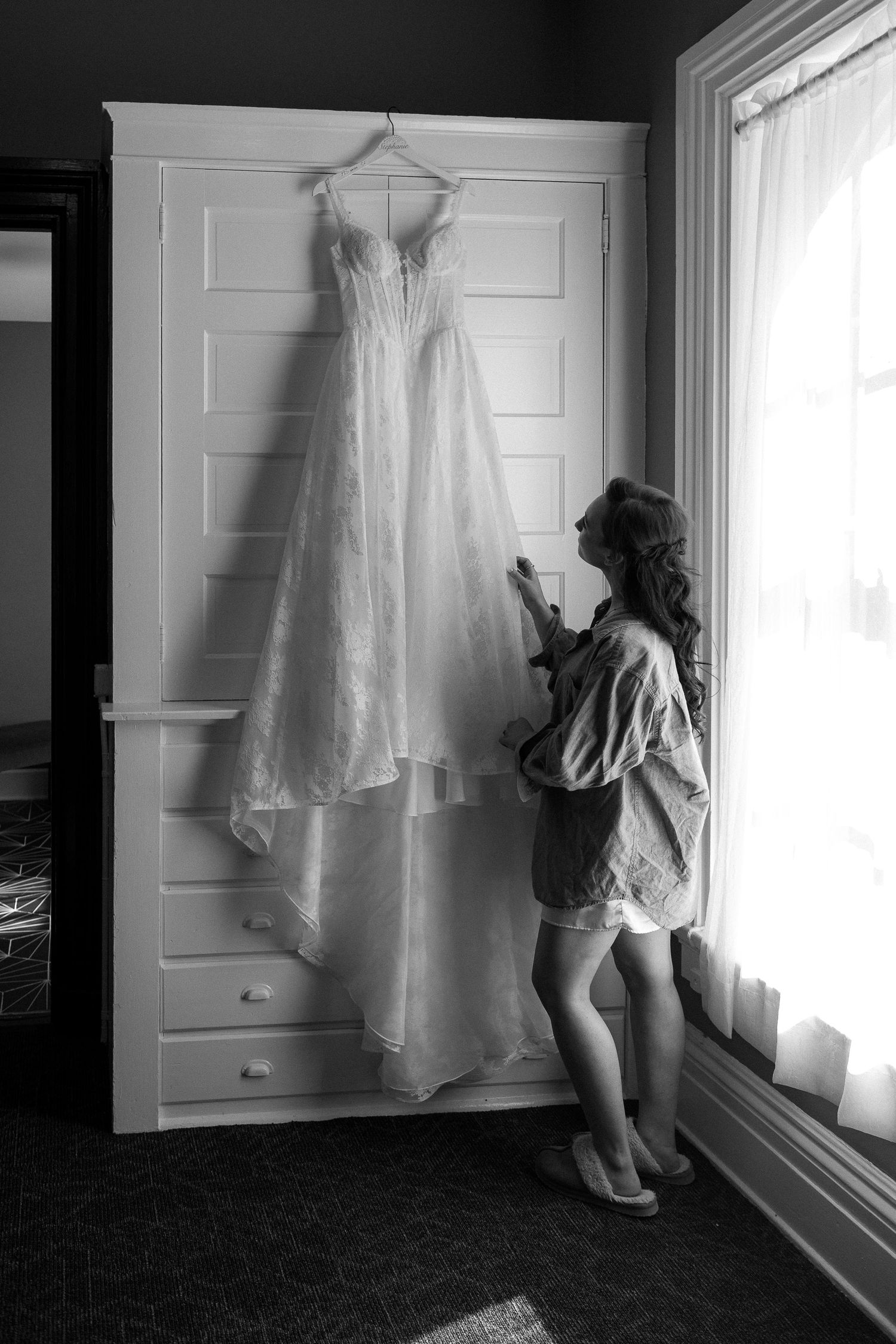 A wedding dress hangs elegantly against a white paneled wall in black and white photography.