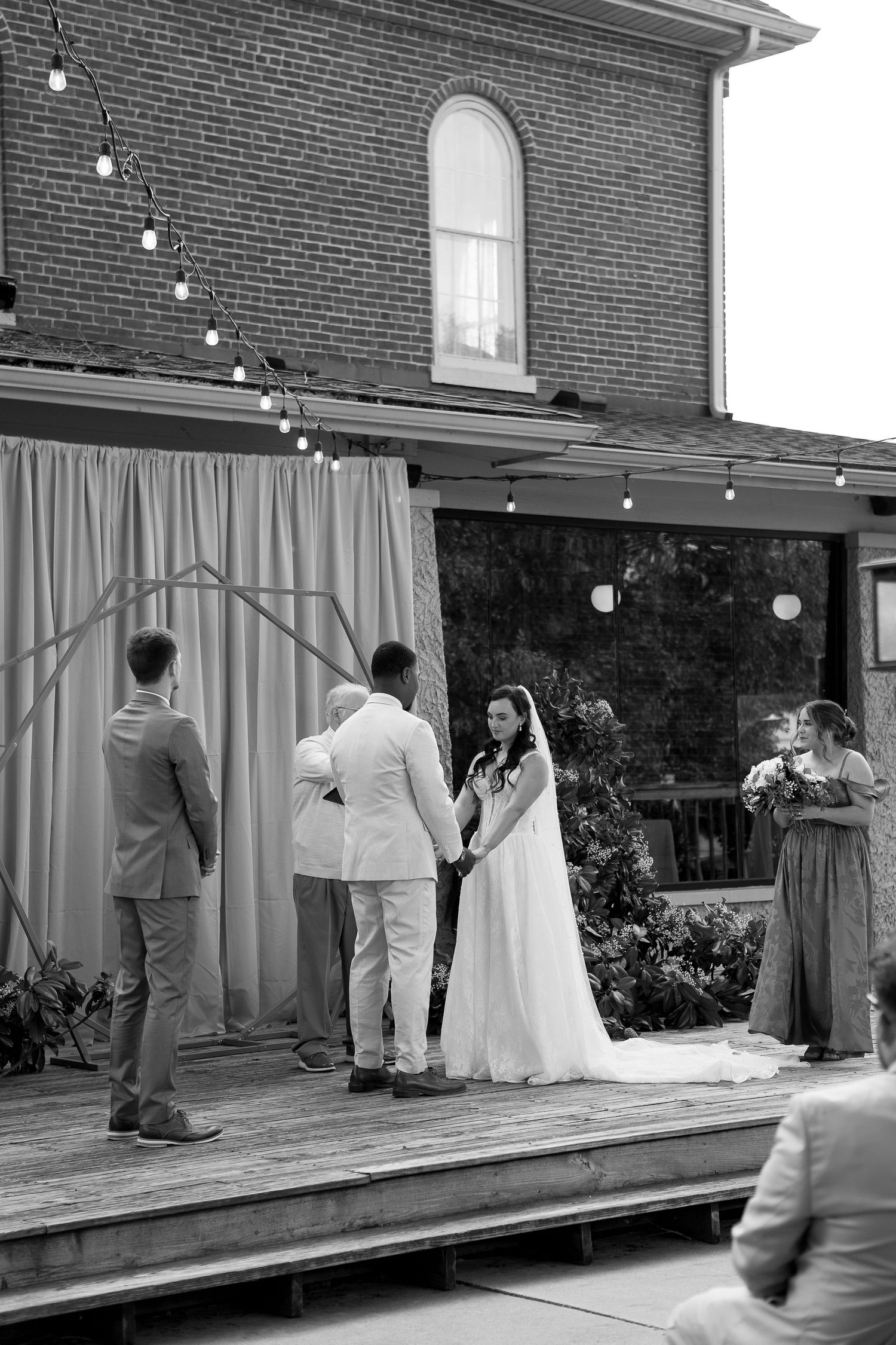 Black and white photo of a wedding ceremony on an outdoor deck with string lights and brick building backdrop.