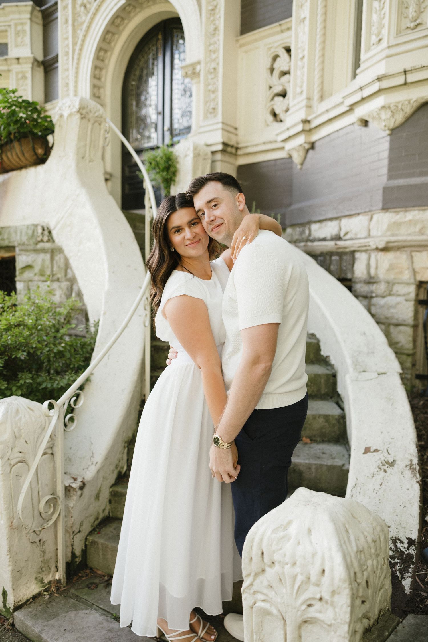 Couple embracing in front of an ornate white staircase of a historic building on a sunny day.