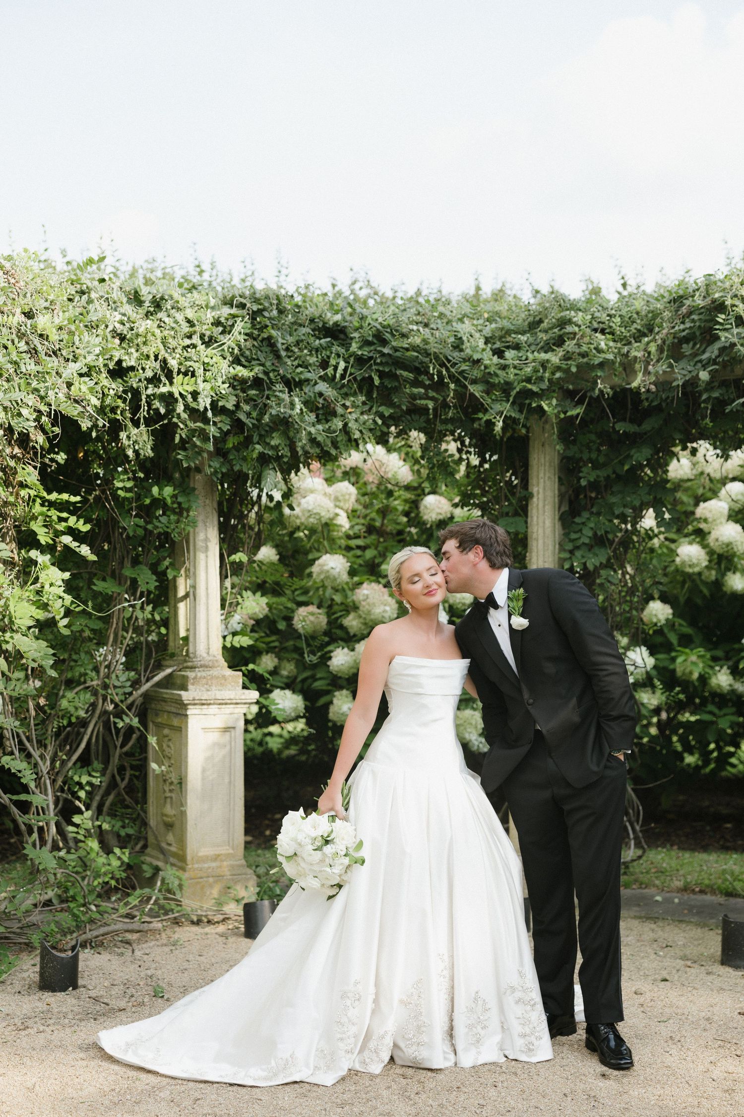 Bride in strapless white gown and groom in black tuxedo share a romantic kiss in a lush garden setting.