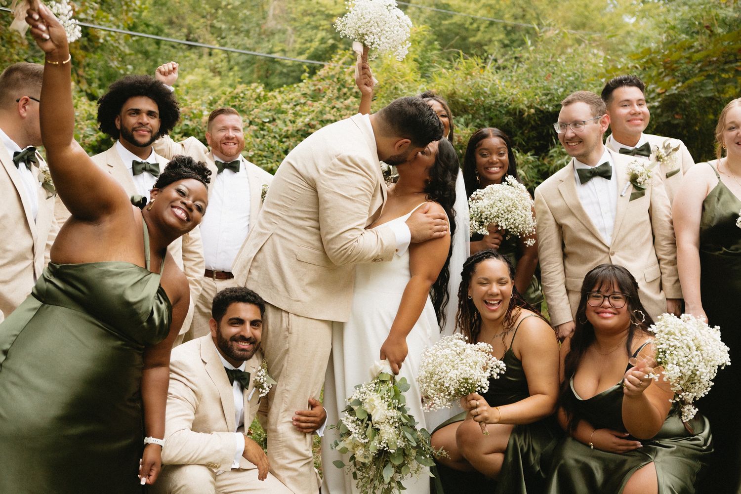 A lively wedding party celebrates outdoors with bouquets and joyful expressions in a natural garden setting.
