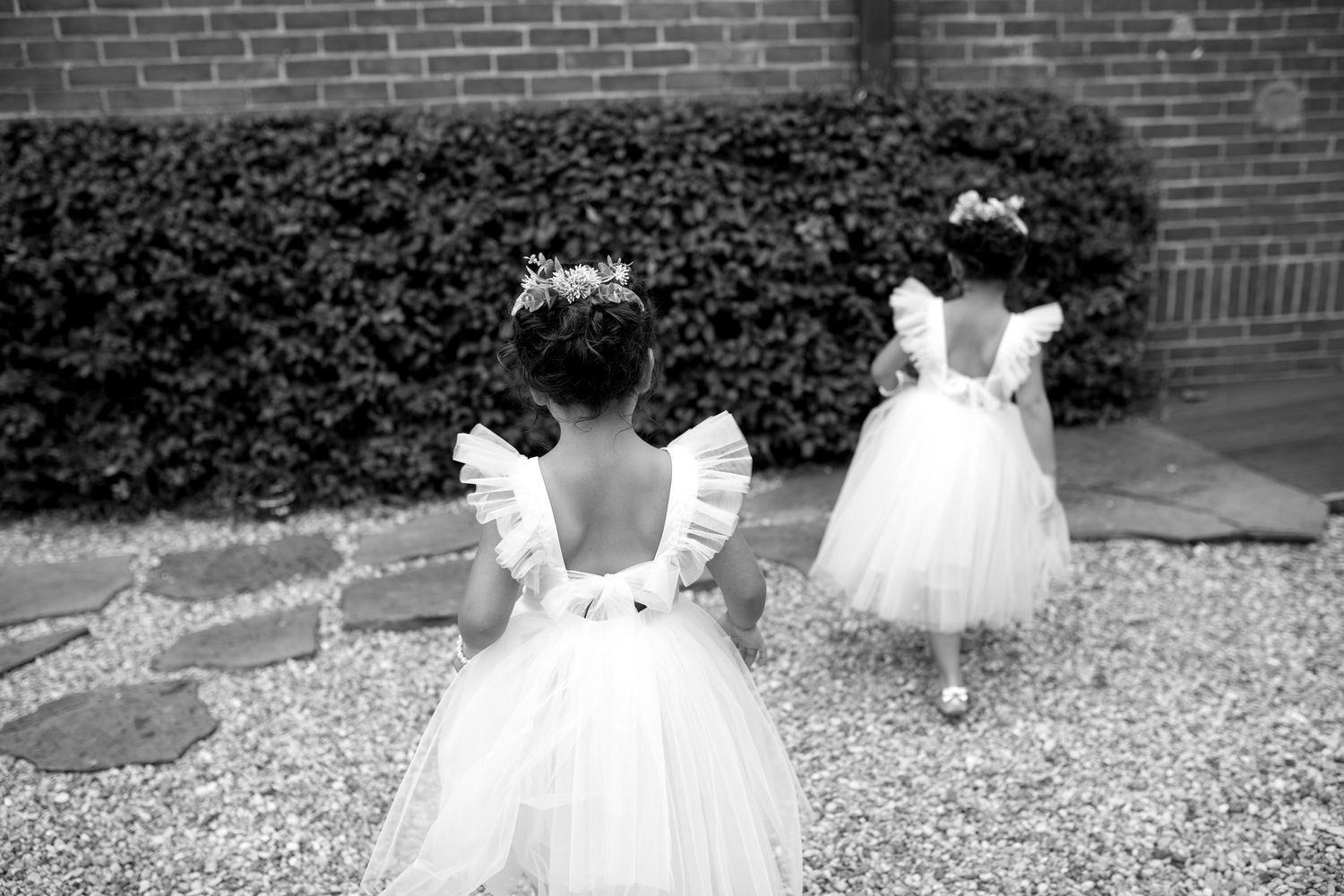 Two young children in white ruffled dresses with open backs stand near a brick wall and hedges in this black and white photo.