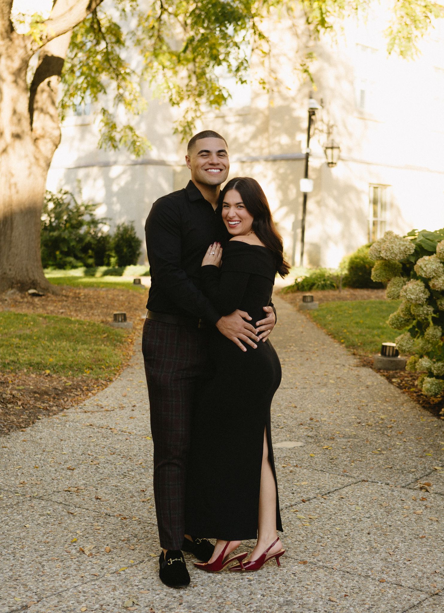 A romantic couple embraces on a garden path with lush greenery and hydrangeas in the background in warm sunlight.