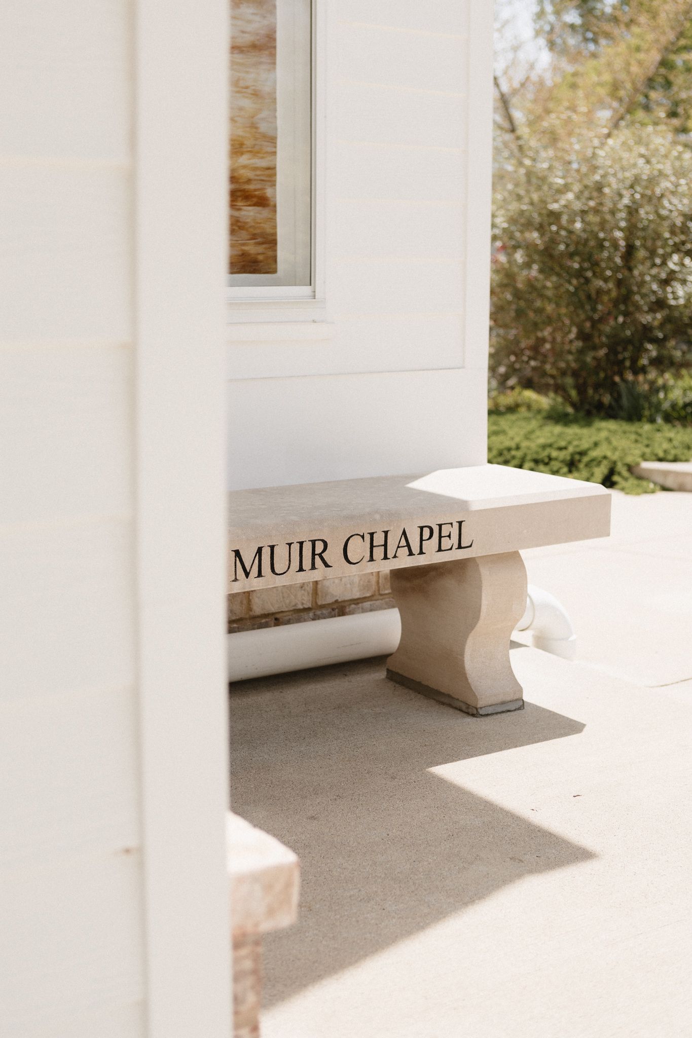 A stone bench inscribed with 'Muir Chapel' sits in a white architectural alcove with natural sunlight and greenery visible.