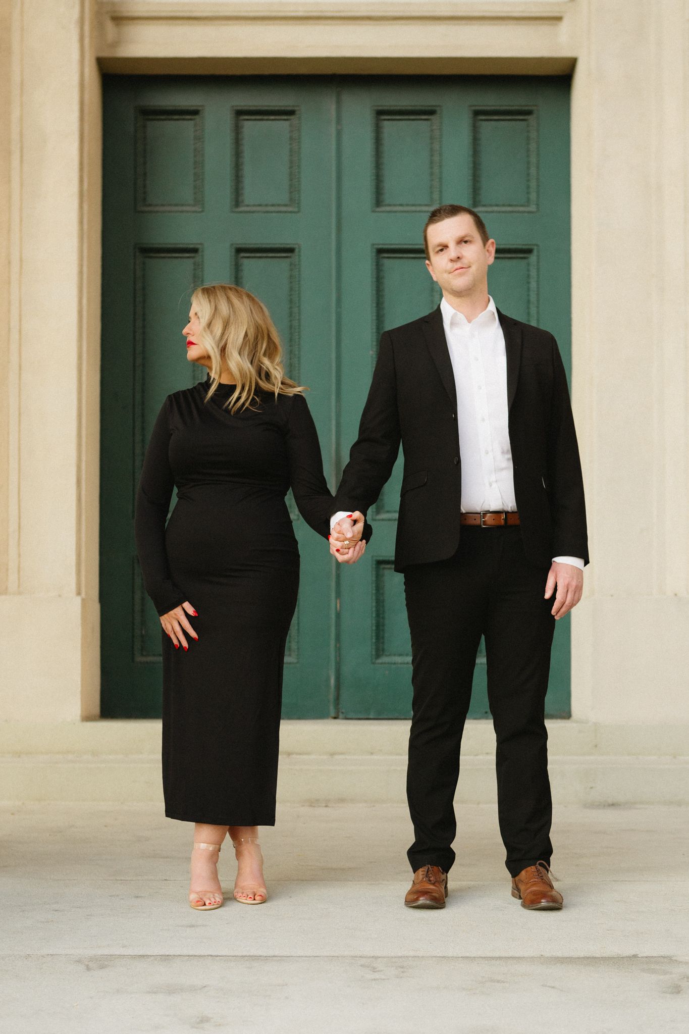 A couple poses in front of a large green door, wearing black formal attire and holding hands.