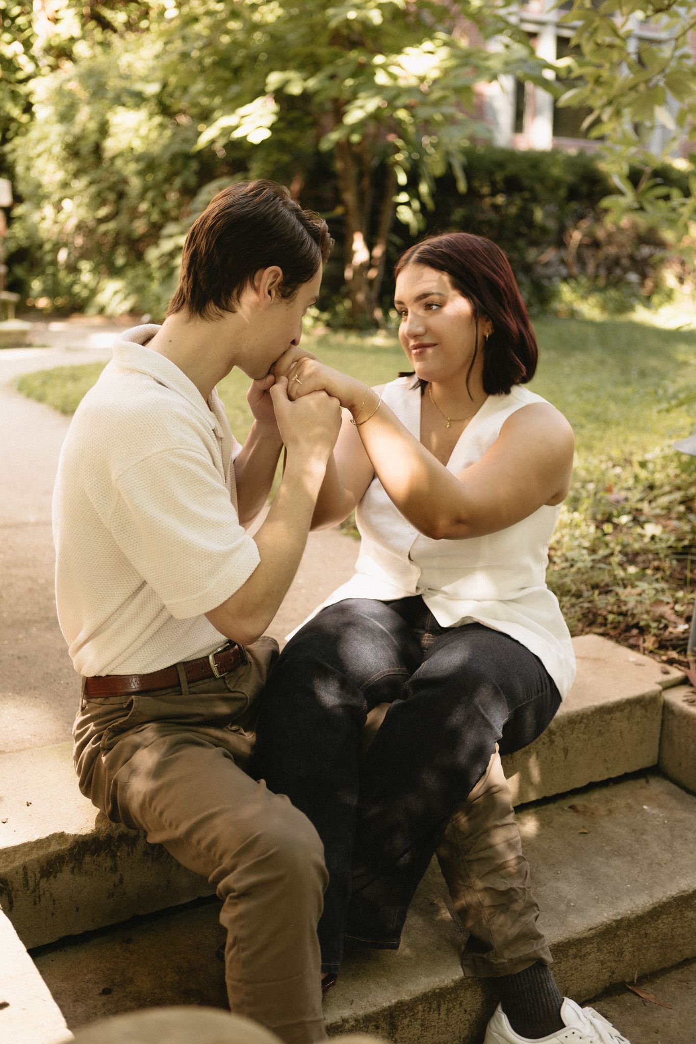 A couple shares an intimate moment sitting together on stone steps in a sunlit garden setting.
