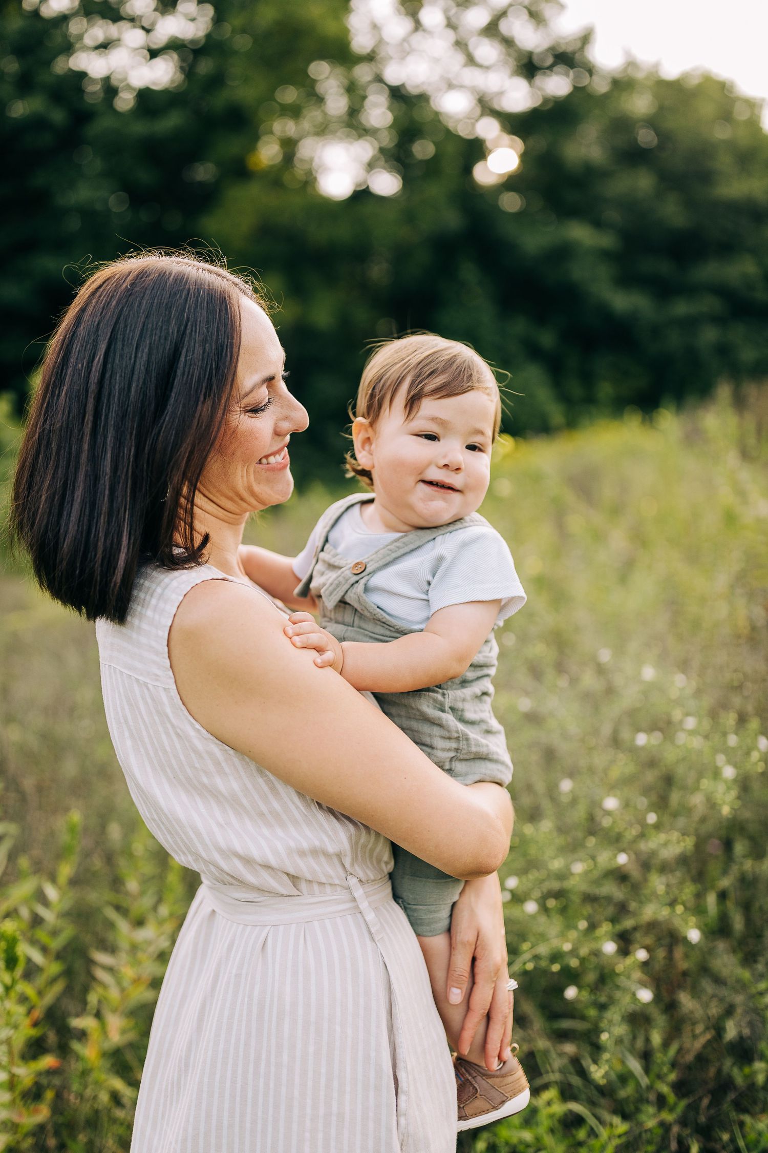 Mom holding her young son in a grassy field with wildflowers during the summertime at sunset.