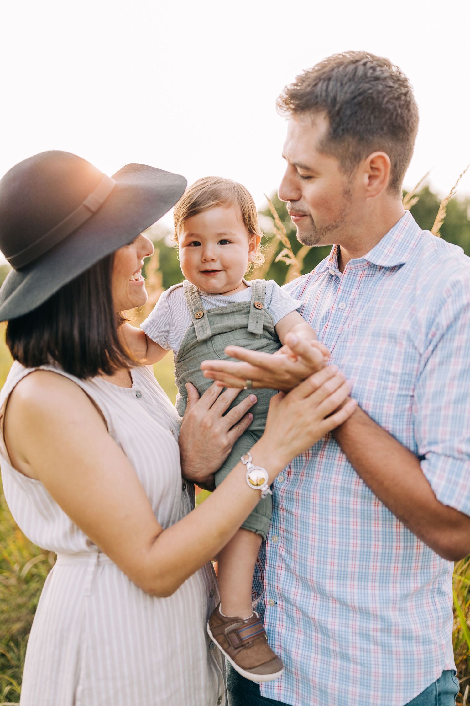 Mom and dad holding their young son in a grassy field during the summertime at sunset.