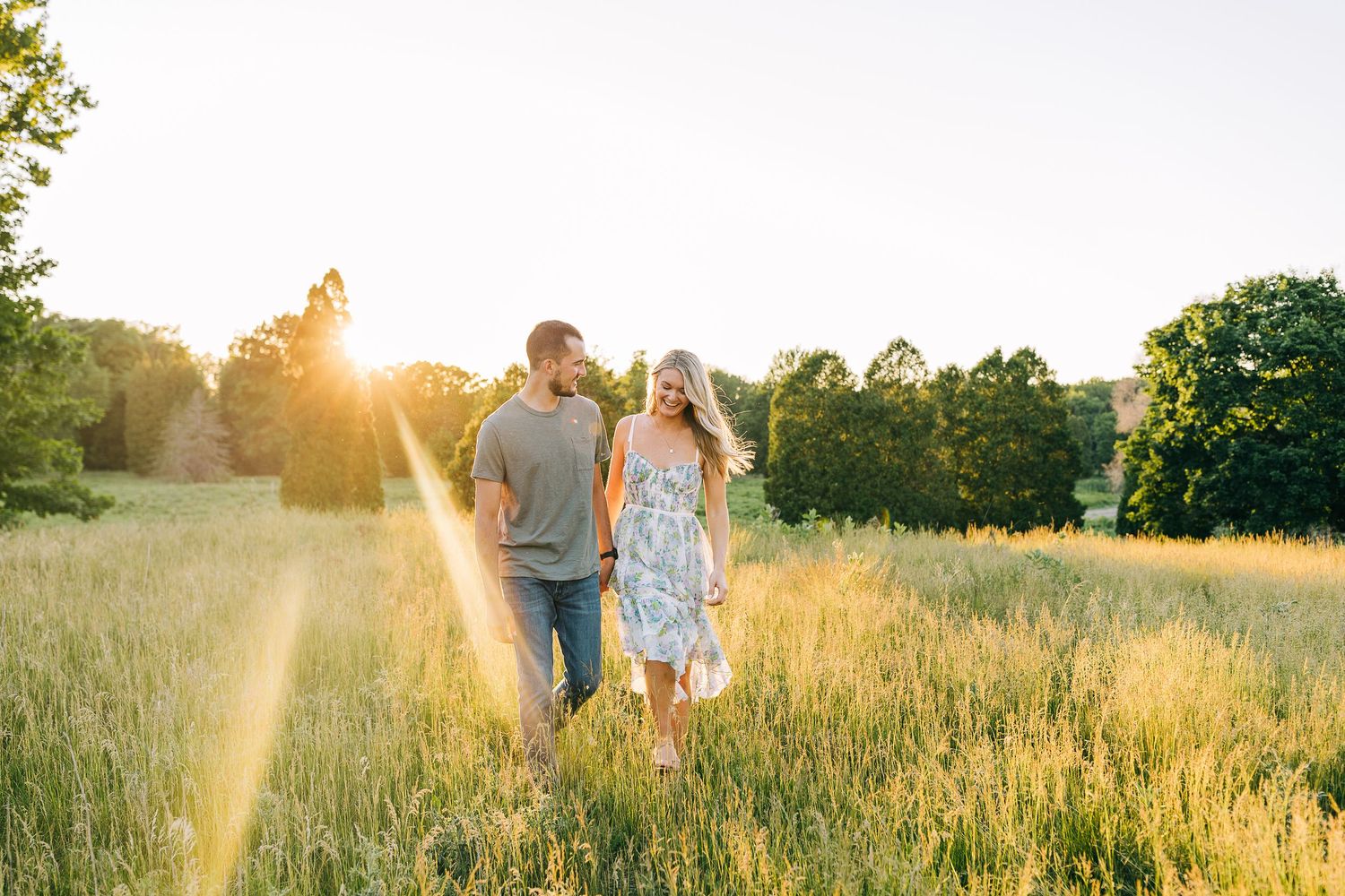 Engaged couple holding hands while walking in a wide open field during sunset.