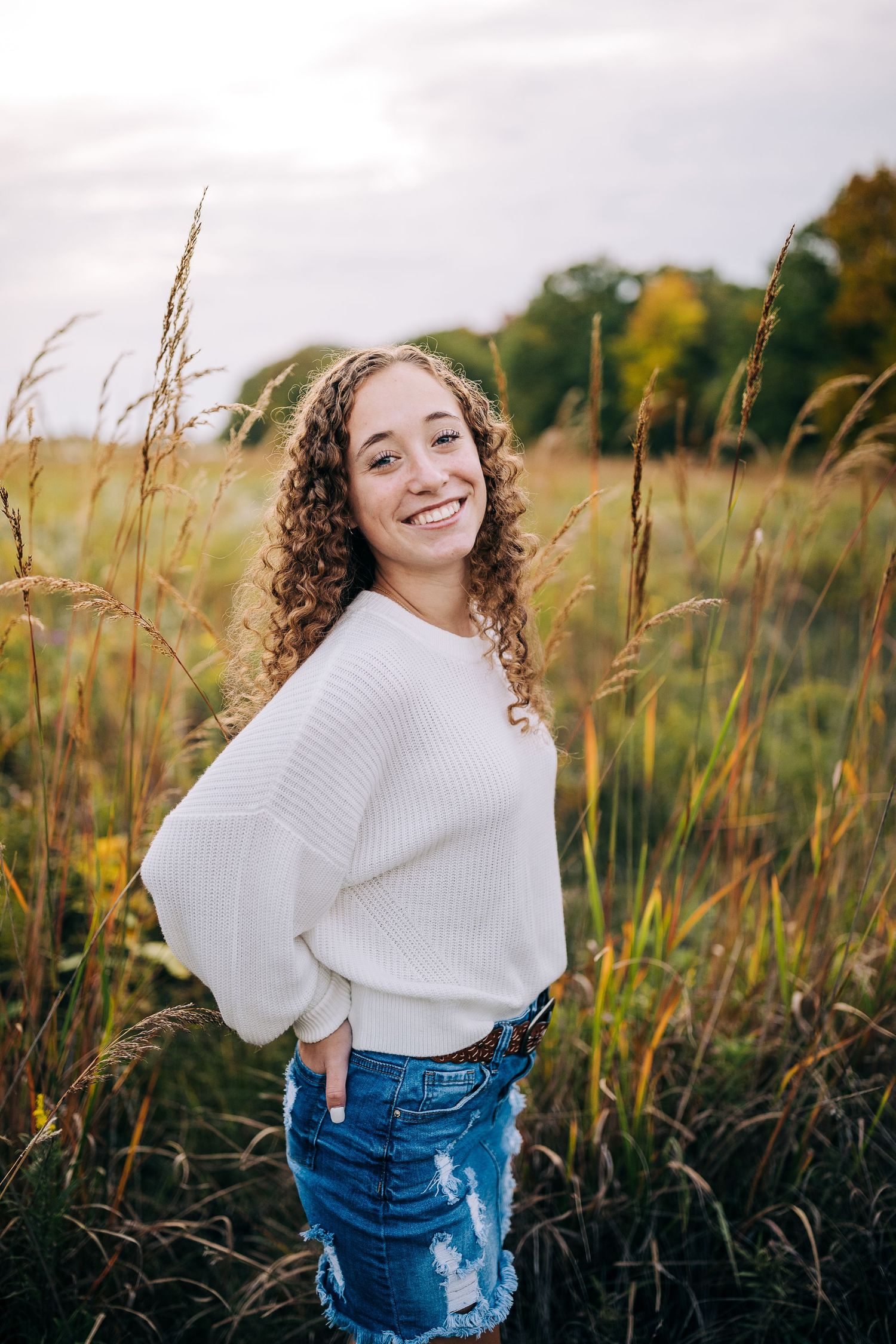 Senior girl wearing a white sweater and jean skirt, standing in a open field in early autumn.