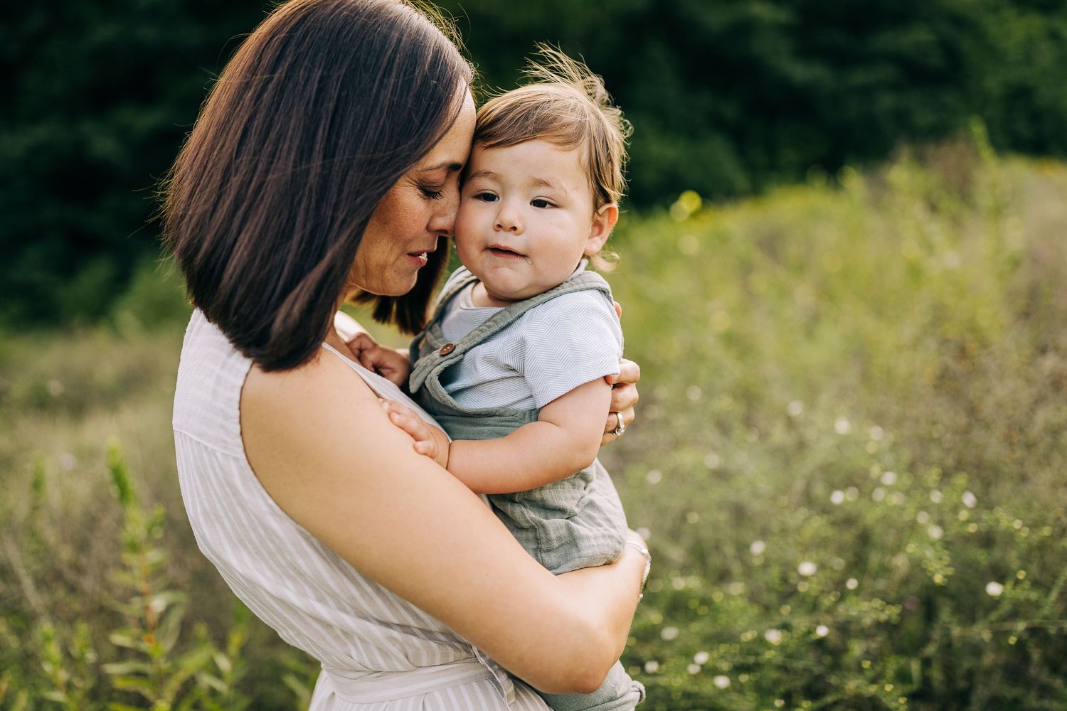 Mom holding her young son in a grassy field with wildflowers during the summertime at sunset.