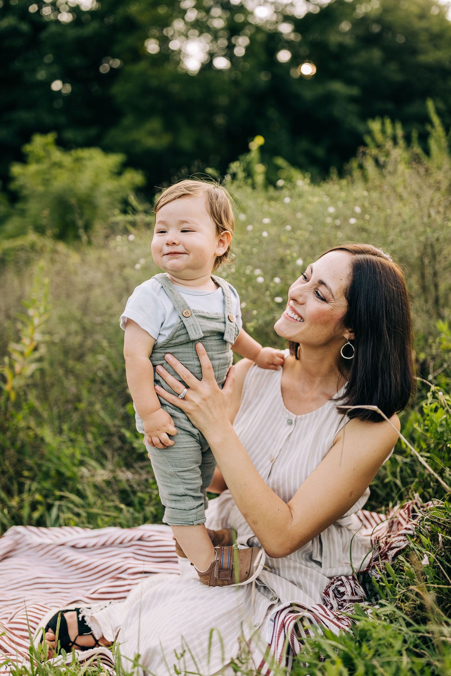 Mom holding her young son in a grassy field with wildflowers during the summertime at sunset.