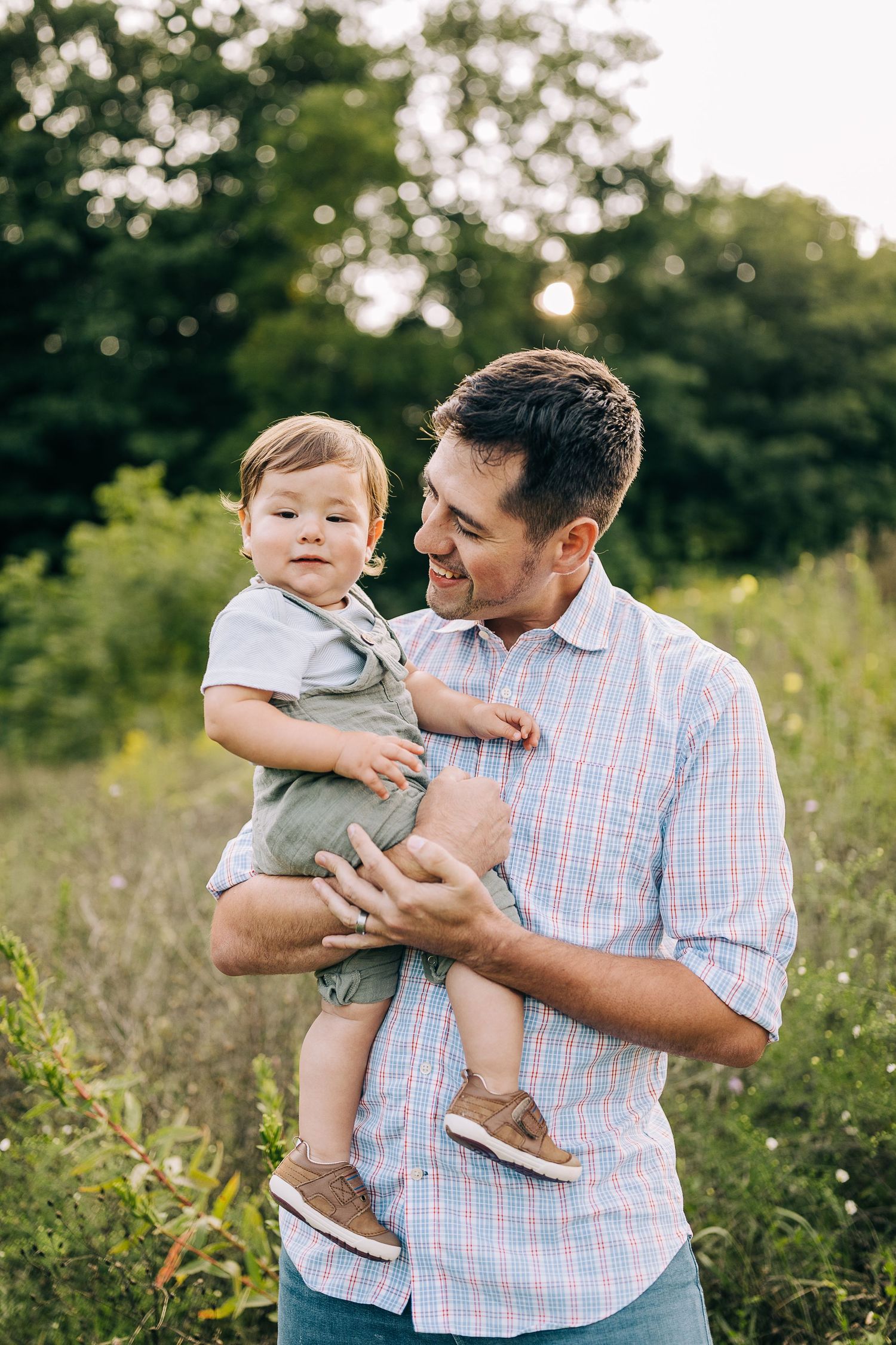 Dad holding her young son in a grassy field with wildflowers during the summertime at sunset.