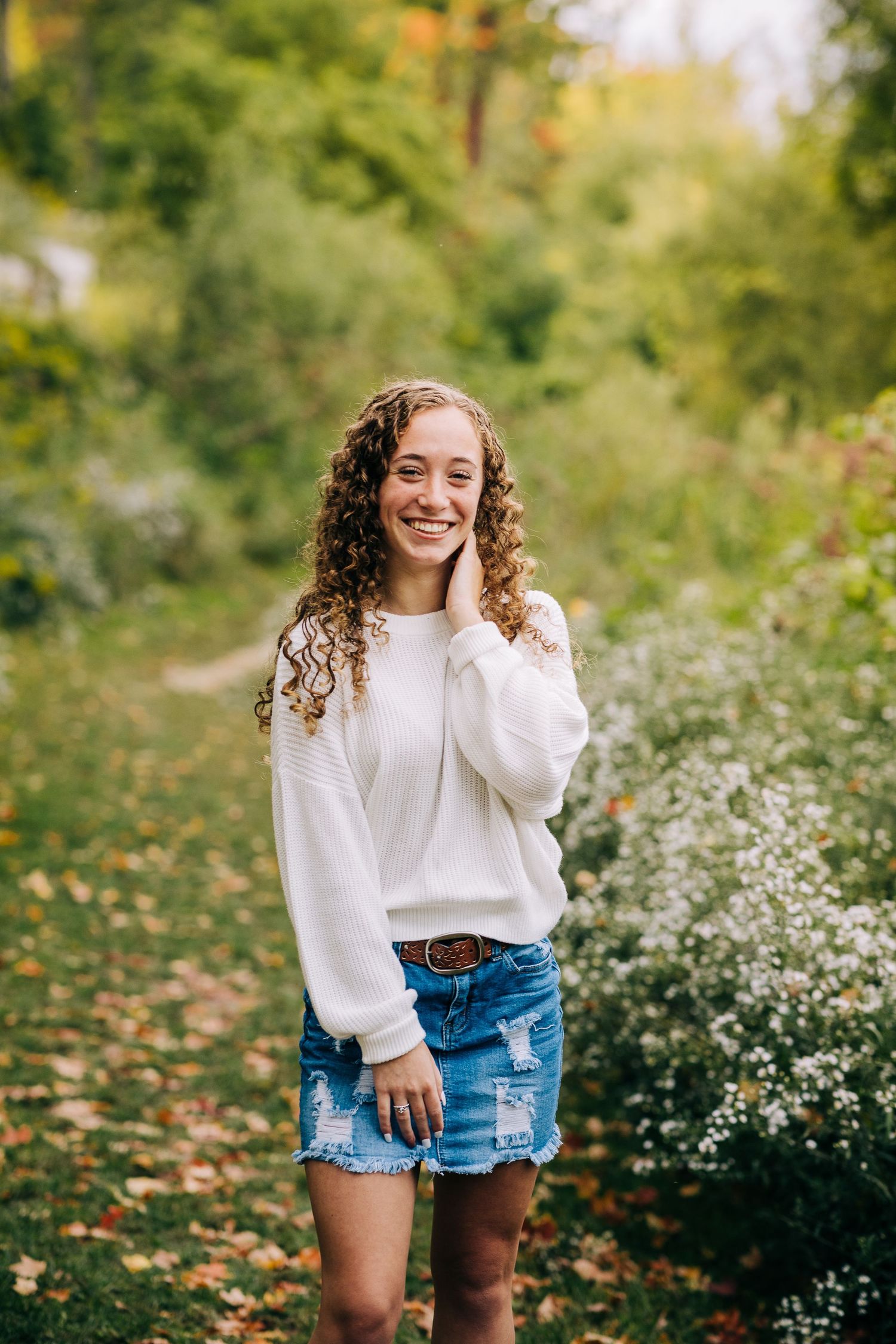 Senior girl wearing a white sweater and jean skirt on a grassy trail in early autumn.