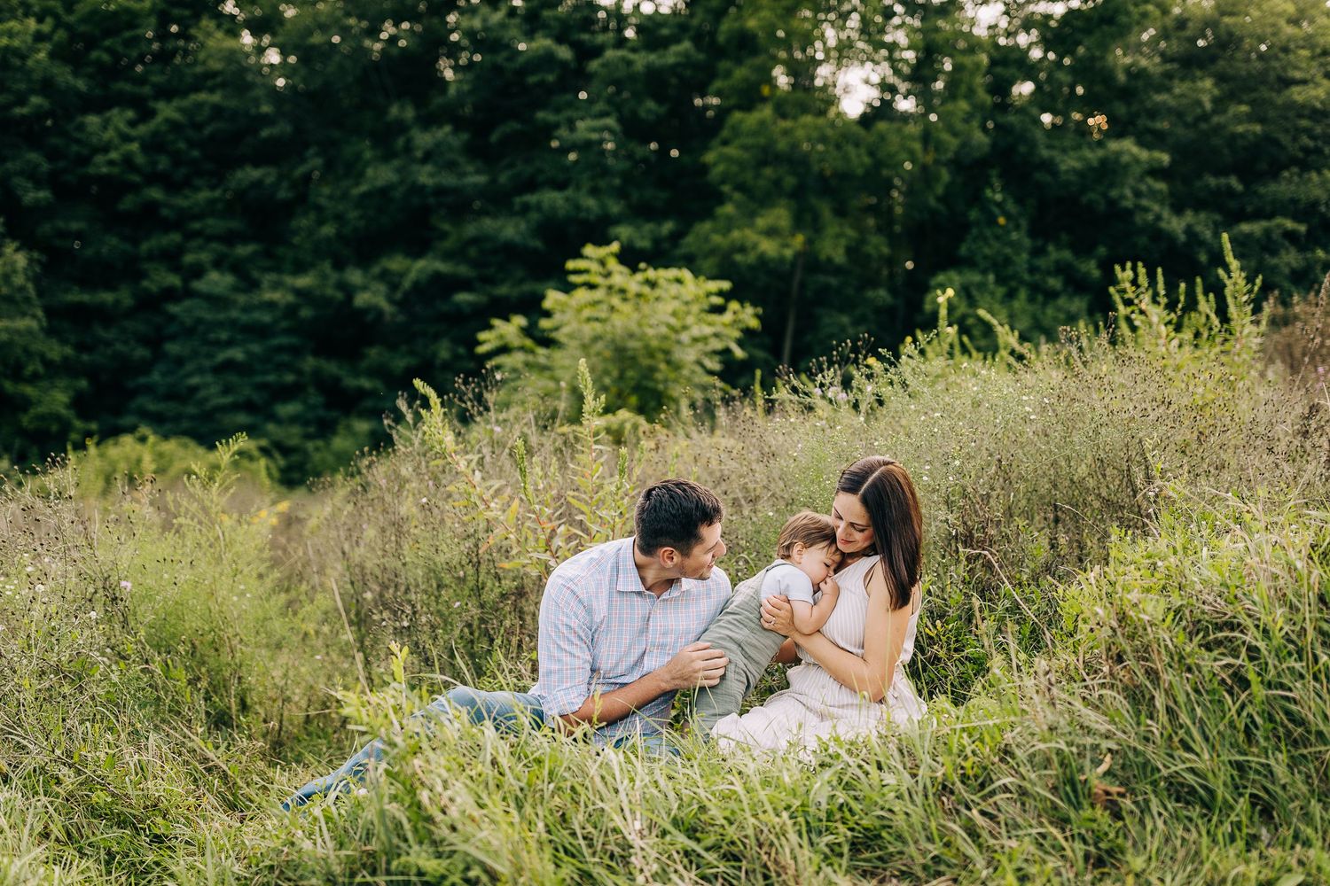 Mom and dad holding their young son in a grassy field during the summertime at sunset.
