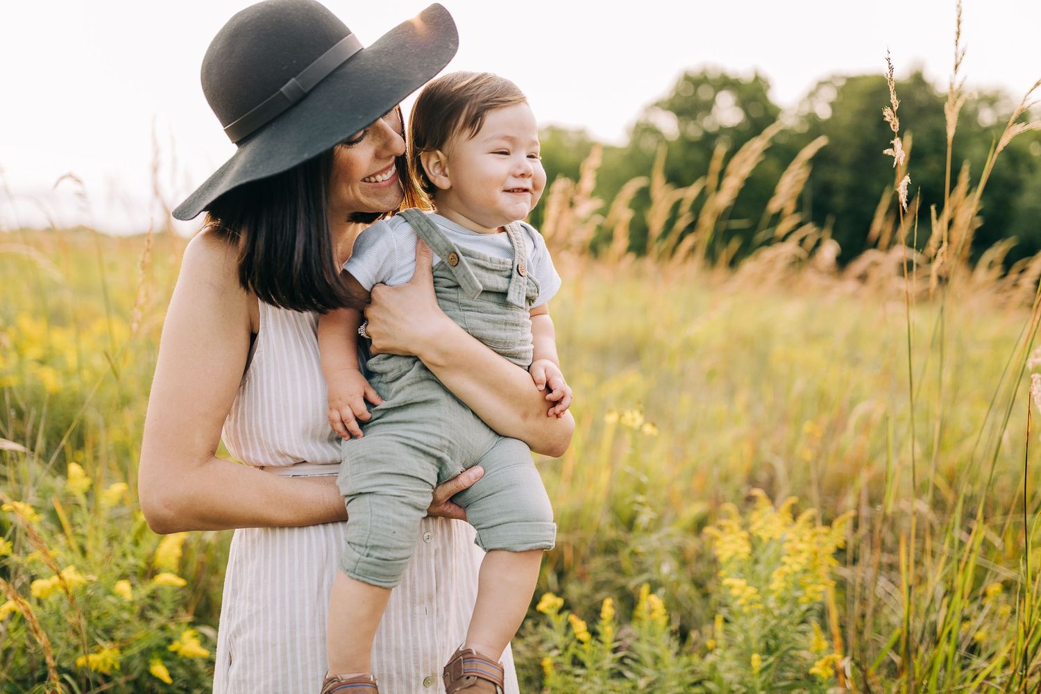 Mom holding her young son in a grassy field with wildflowers during the summertime at sunset.