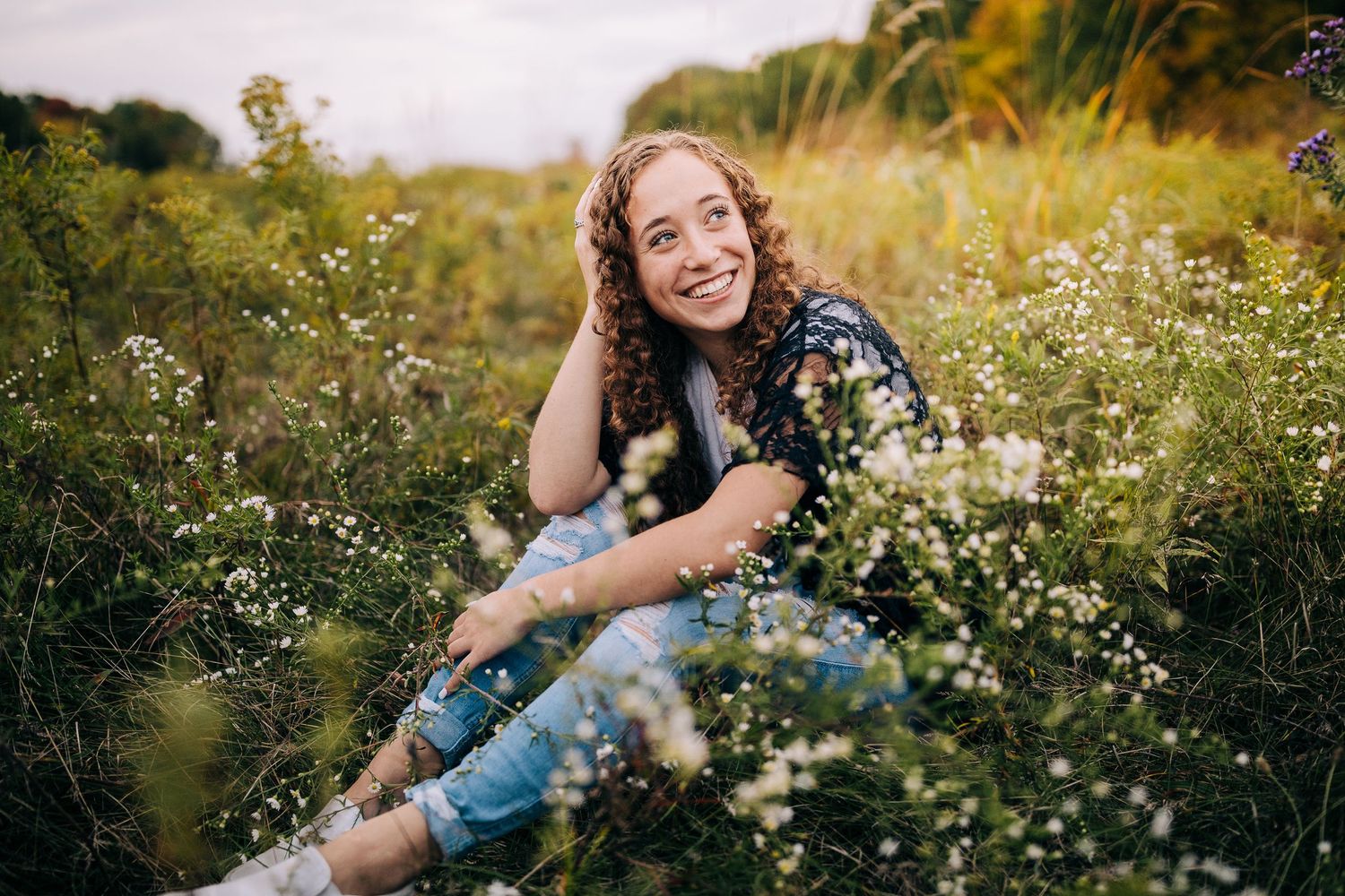 Senior girl wearing a lacy black sweater and jeans, sitting in a field of wildflowers in early autumn.