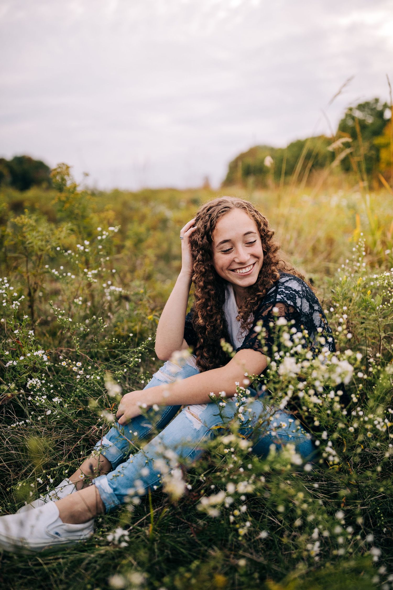 Senior girl wearing a lacy black sweater and jeans, sitting in a field of wildflowers in early autumn.