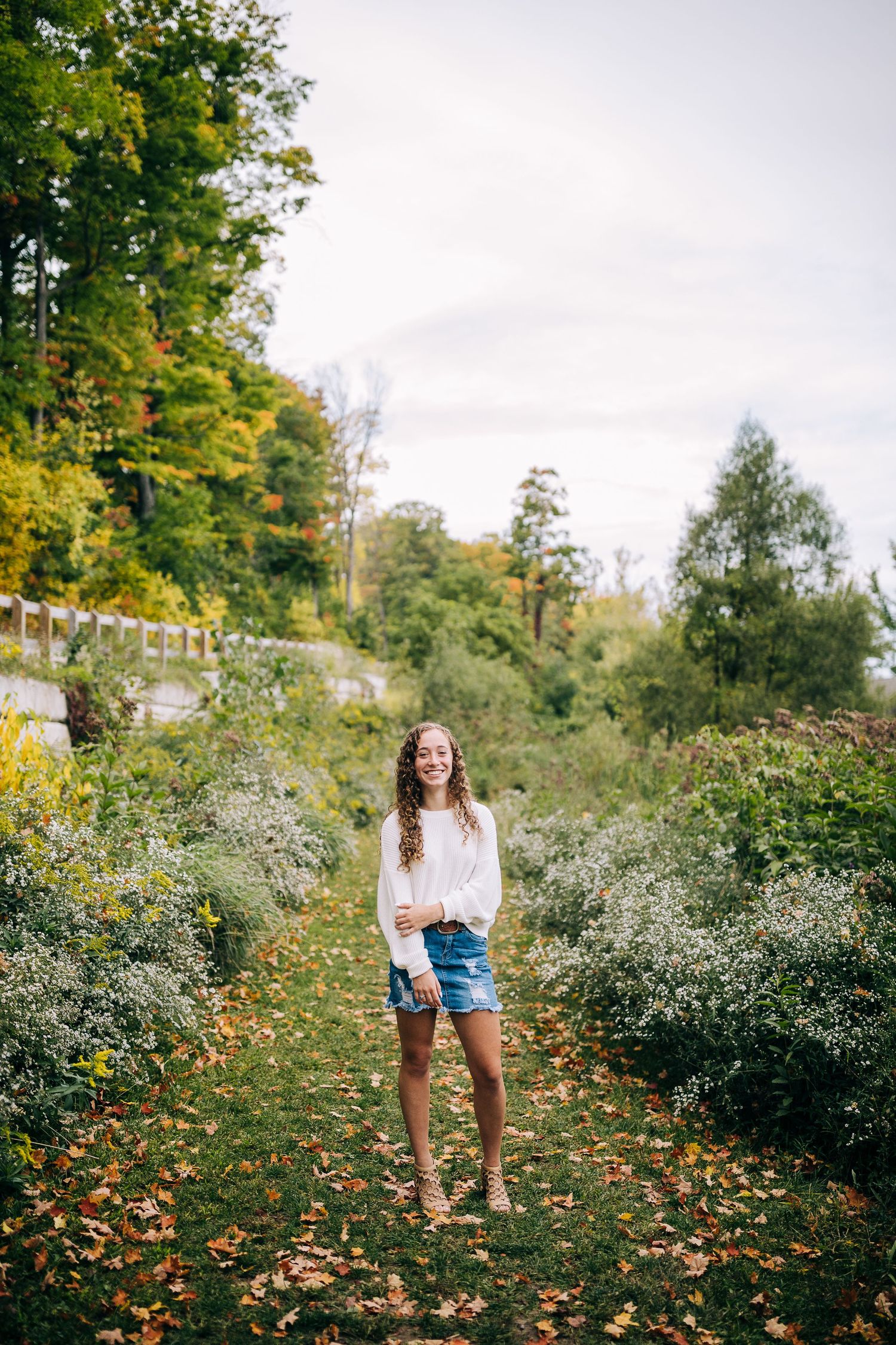 Senior girl wearing a white sweater and jean skirt on a grassy trail in early autumn.