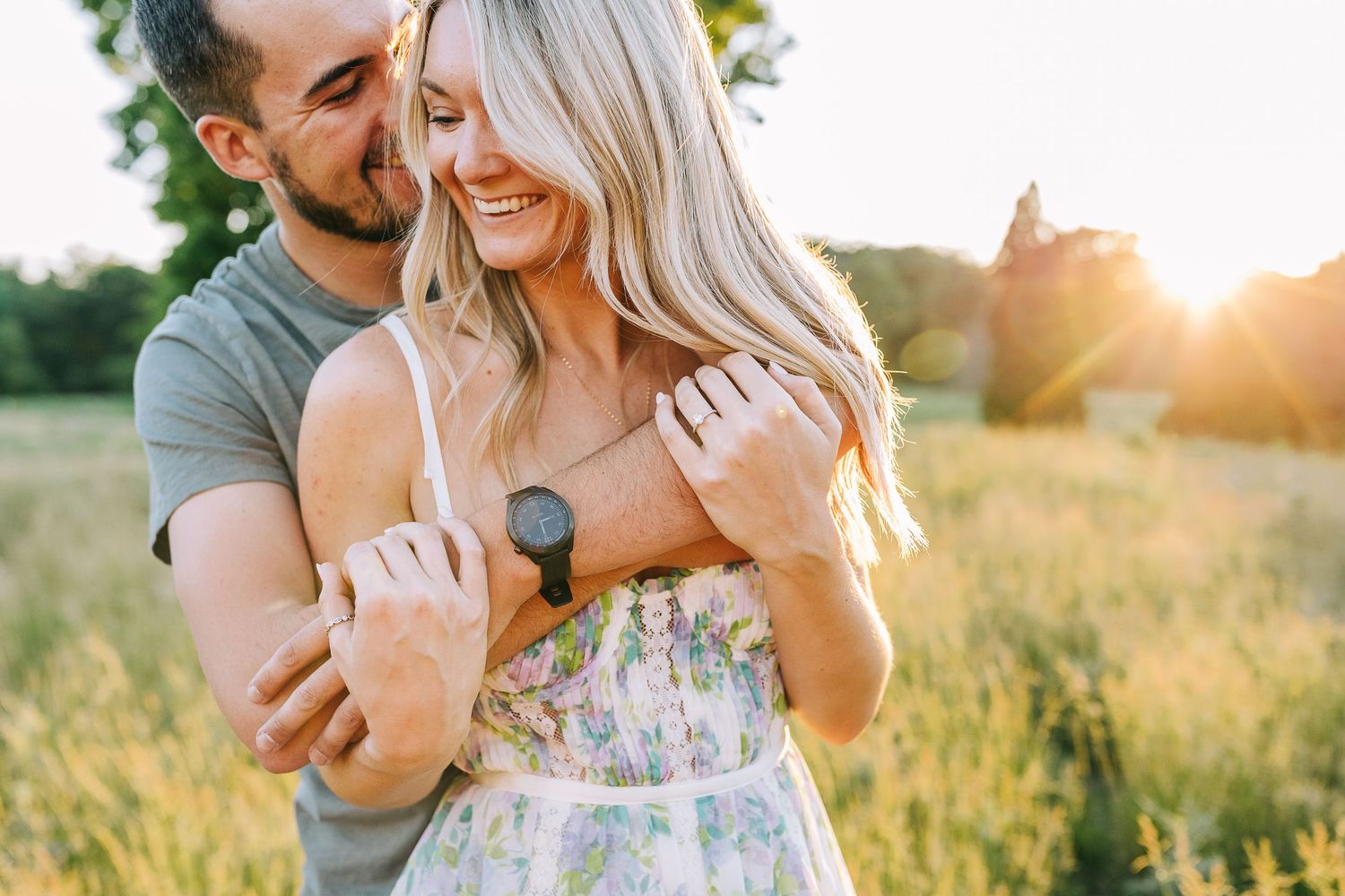 Engaged couple sharing a warm embrace in a wide open field during sunset.