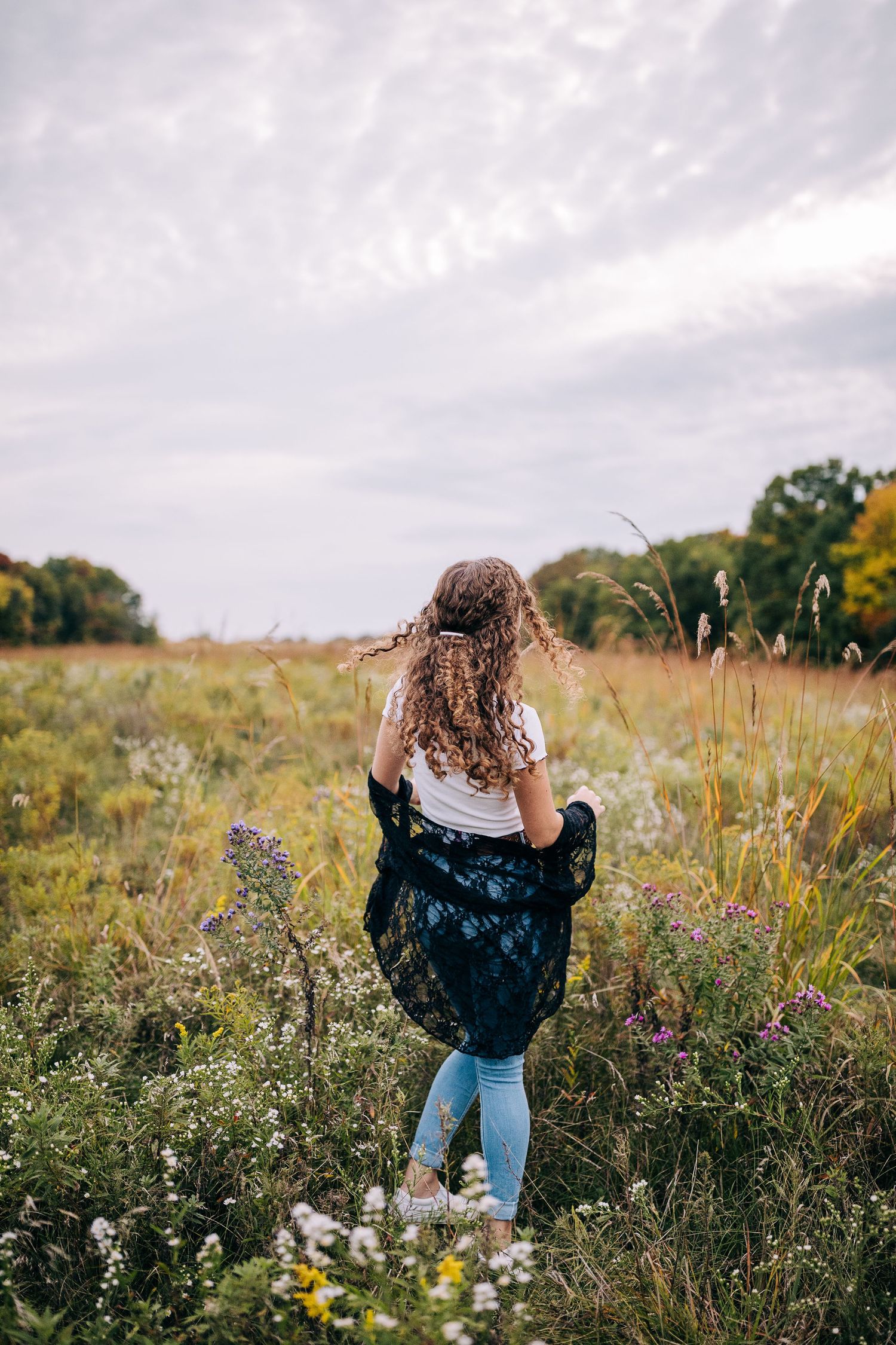 Senior girl wearing a lacy black sweater and jeans, standing  in a field of wildflowers in early autumn.