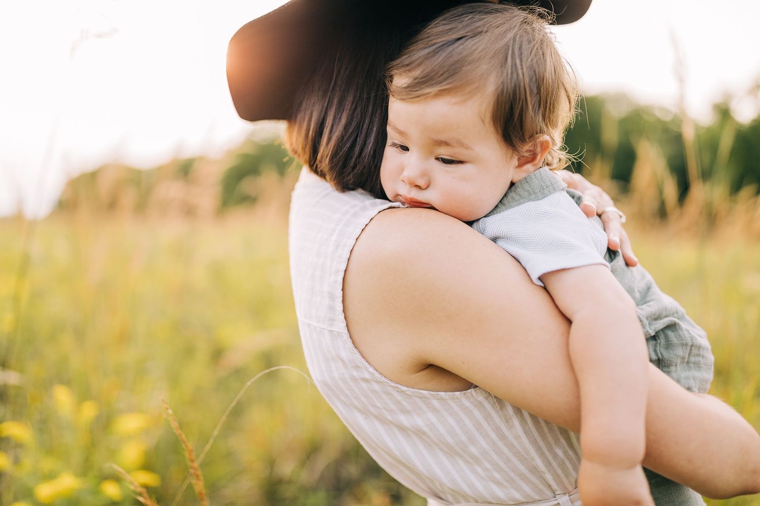 Mom holding her young son in a grassy field with wildflowers during the summertime at sunset.