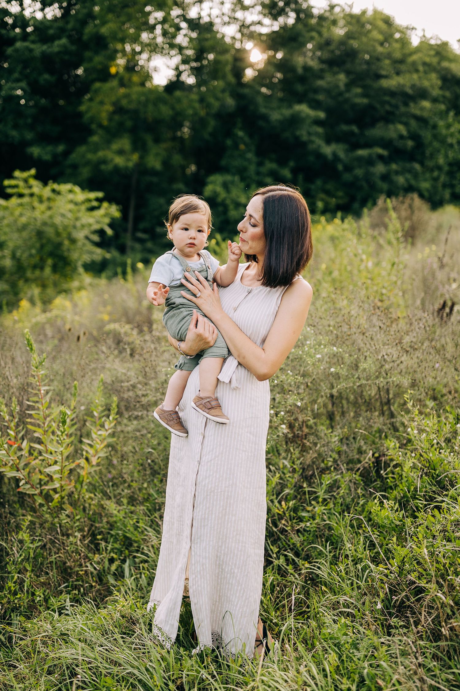 Mom holding her young son in a grassy field with wildflowers during the summertime at sunset.
