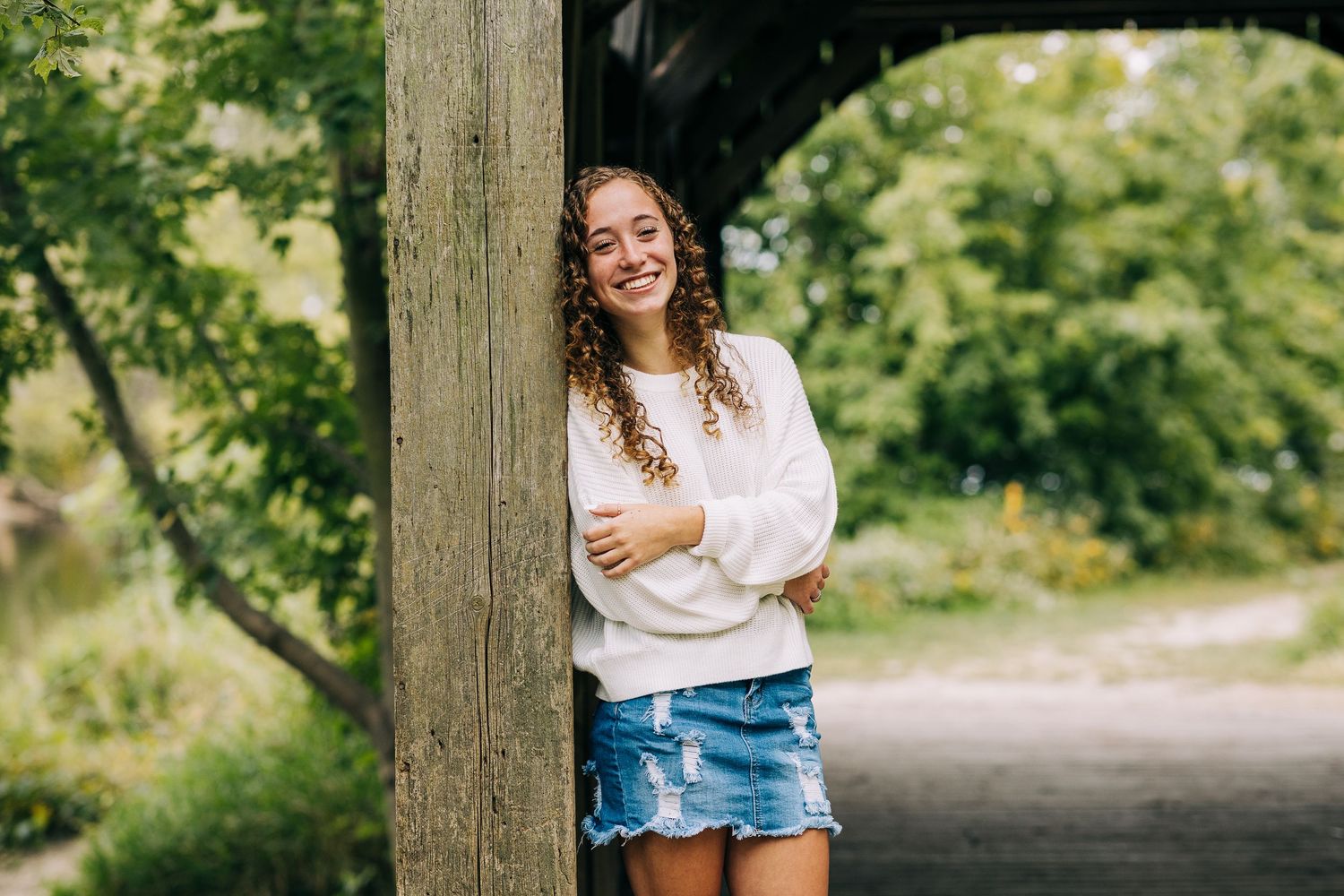 Senior girl wearing a white sweater and jean skirt while standing against a covered wooden bridge in early autumn.