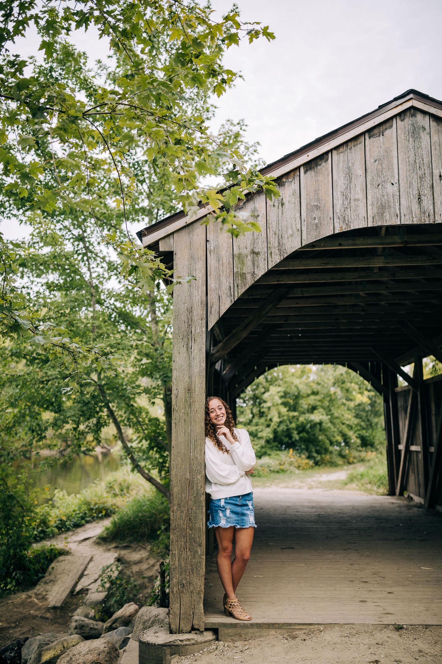 Senior girl wearing a white sweater and jean skirt while standing against a covered wooden bridge in early autumn.