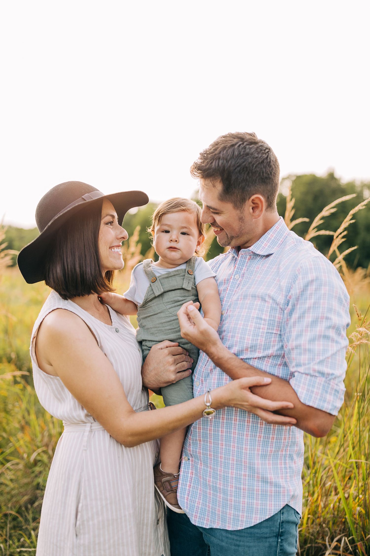 Mom and dad holding their young son in a grassy field during the summertime at sunset.