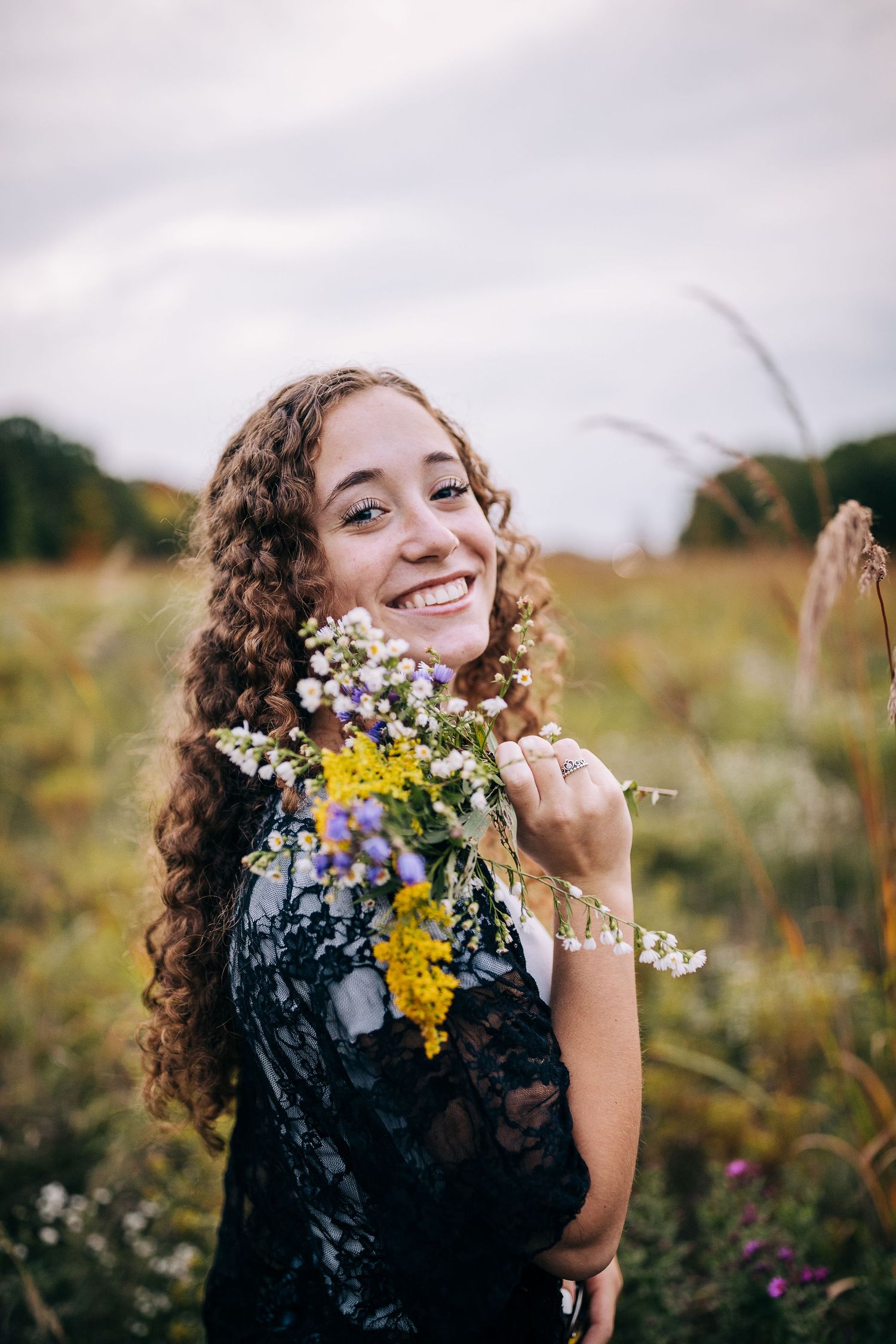 Senior girl wearing a lacy black sweater and jeans, holding a bouquet of wildflowers, while standing in a open field in early autumn.