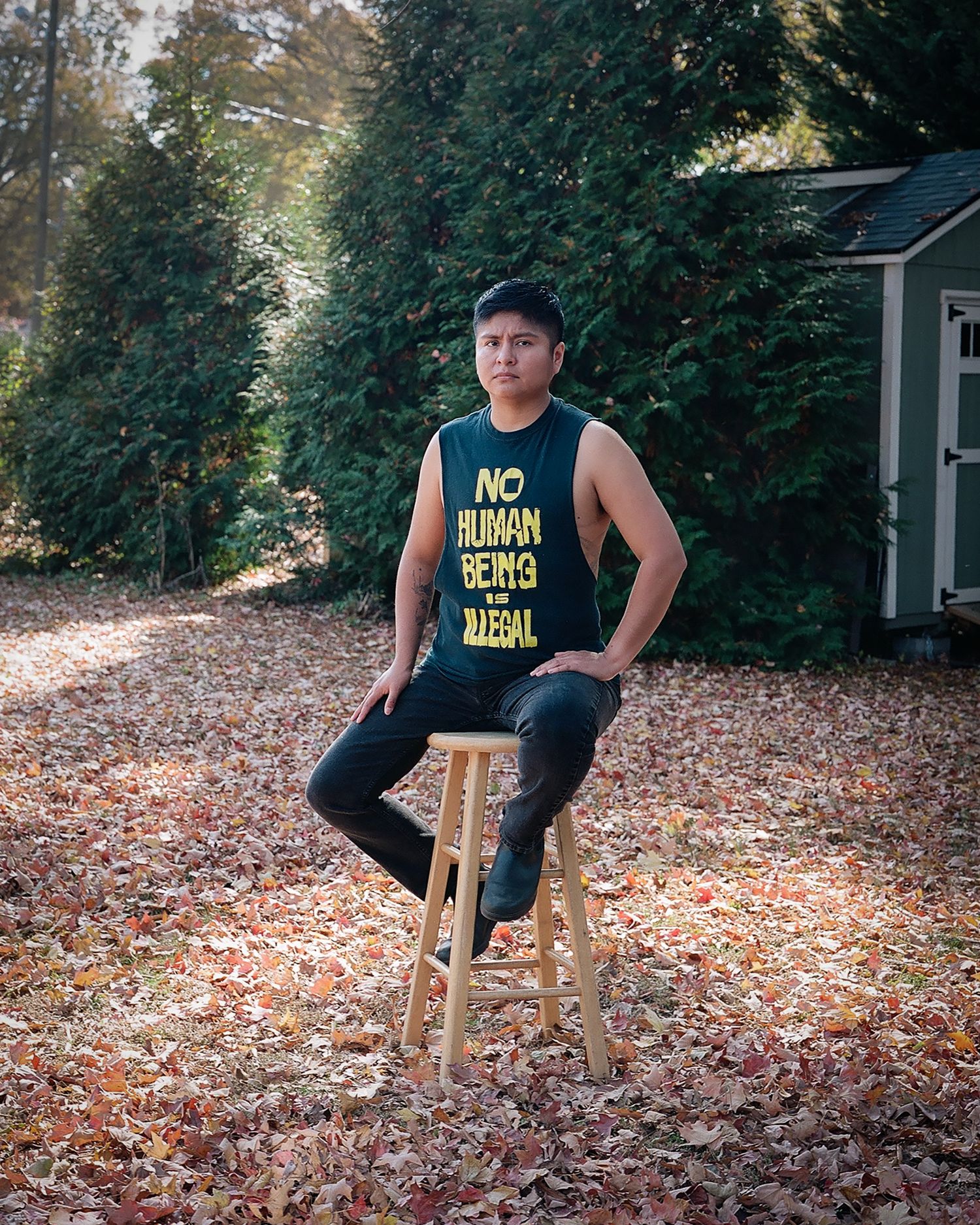 Portrait of a genderqueer latinx sitting on a stool in a yard with a black shirt with yellow letters that reads no human being is illegal.