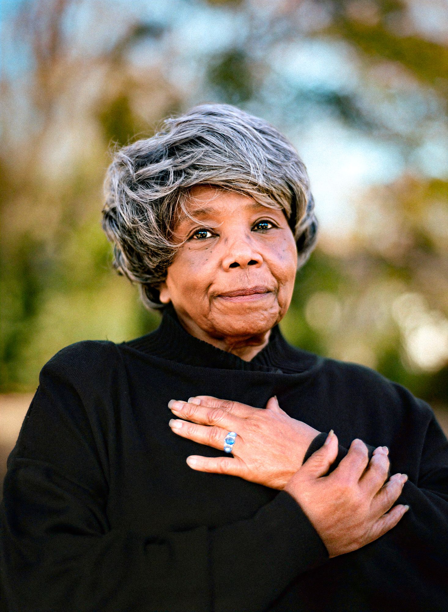 Ms. Peggie, an older Black woman, looks directly at the camera with a gentle smile, her hands crossed over her heart.