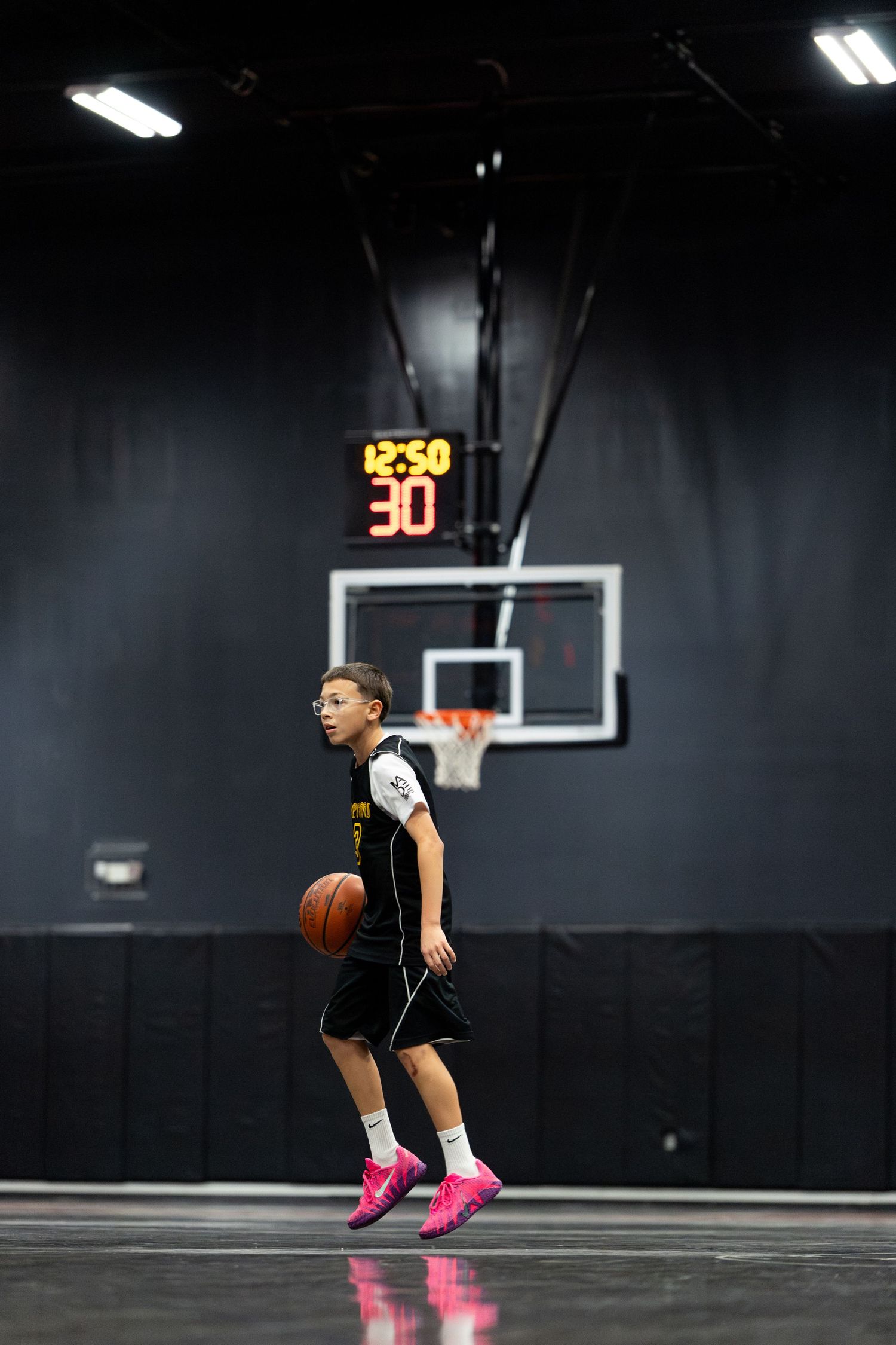 Young basketball player in black uniform dribbling ball mid-jump on indoor court with scoreboard showing 30.
