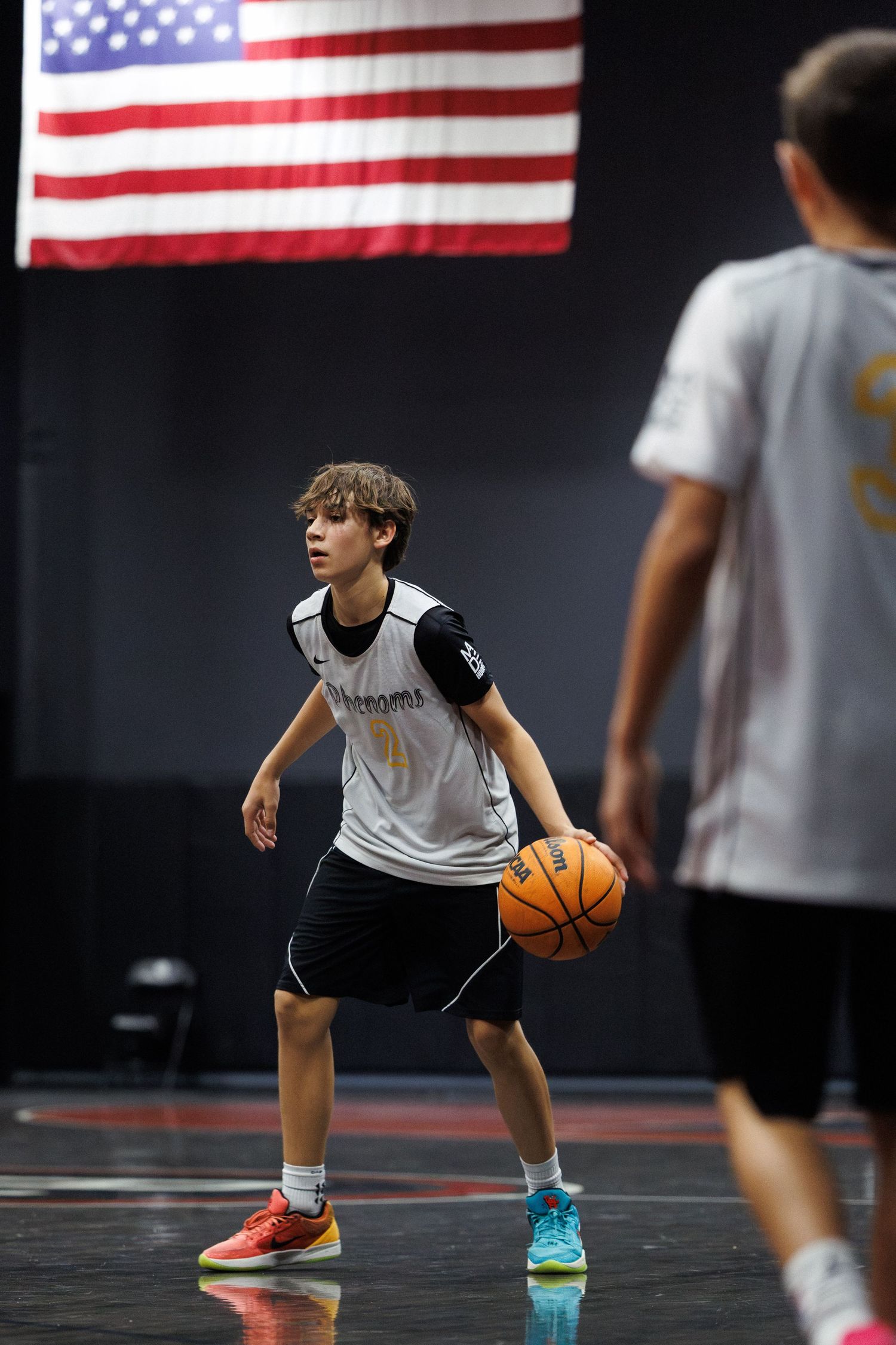 Young male basketball player in white Phenoms jersey dribbling ball on indoor court beneath American flag.