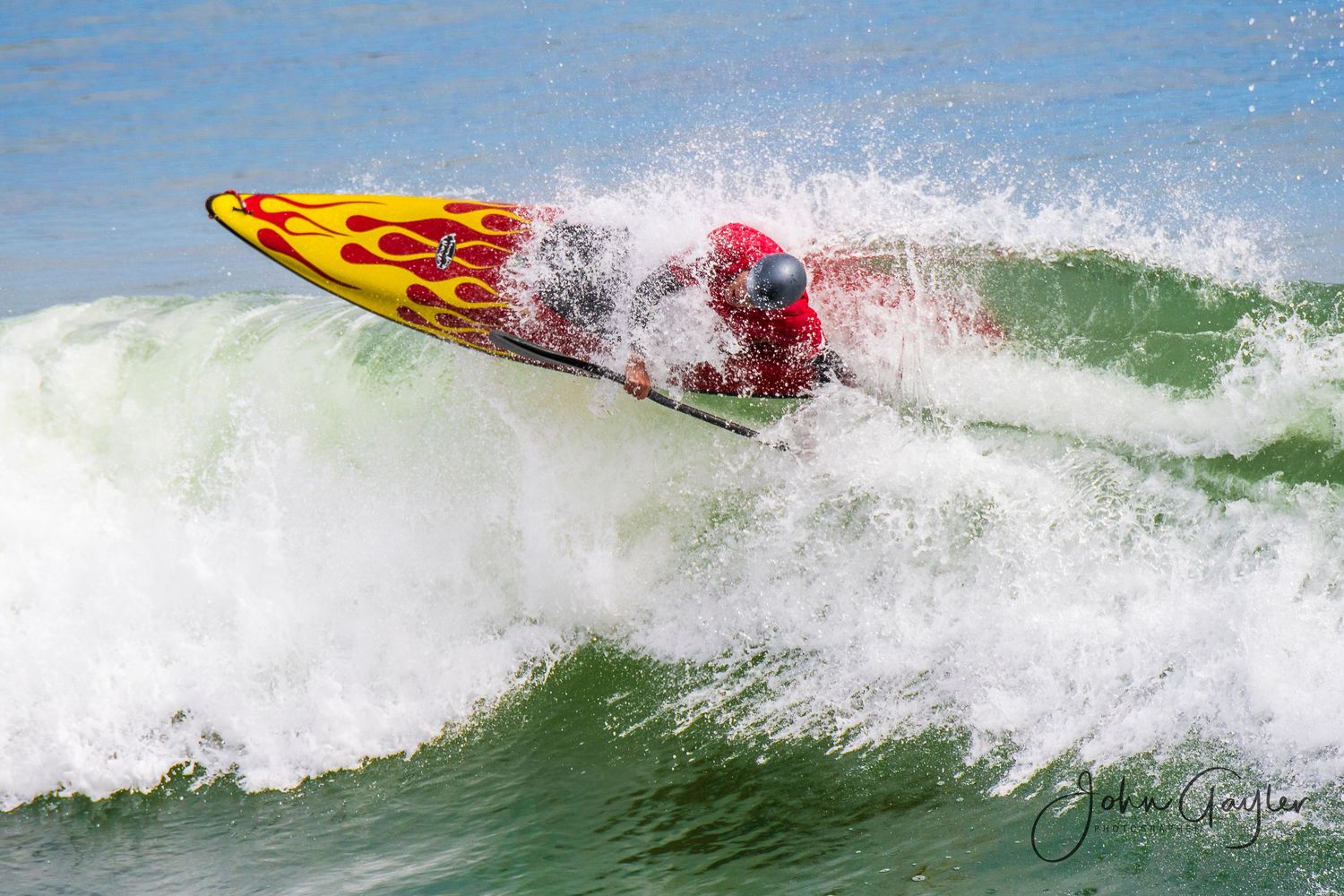 An expert kayak surfer crests a wave in a red surf kayak with yellow flames, at a world surf kayaking championship in Santa Cruz, California