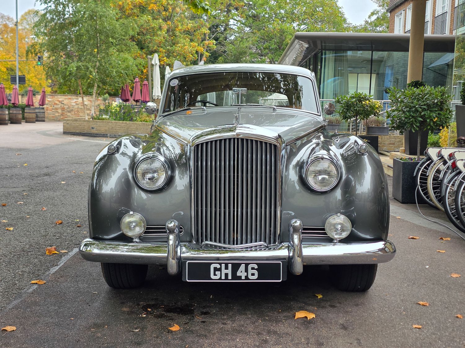 Classic silver Bentley luxury car parked on autumn street with fallen leaves visible on ground.