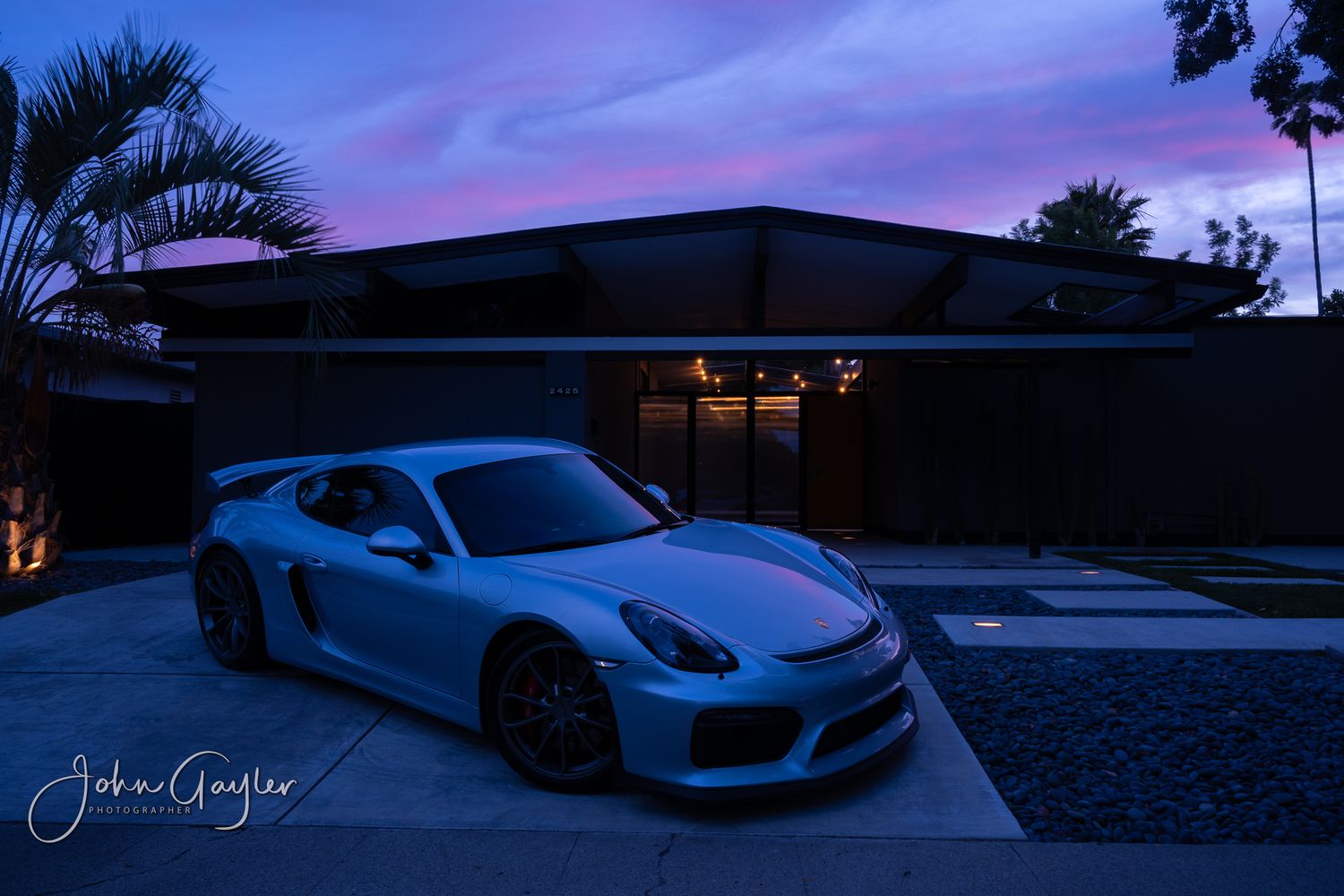 A portrait of a Porsche Carrera GT4 rests outside a modern Eichler designed house with blue pink colors of sunset in the sky