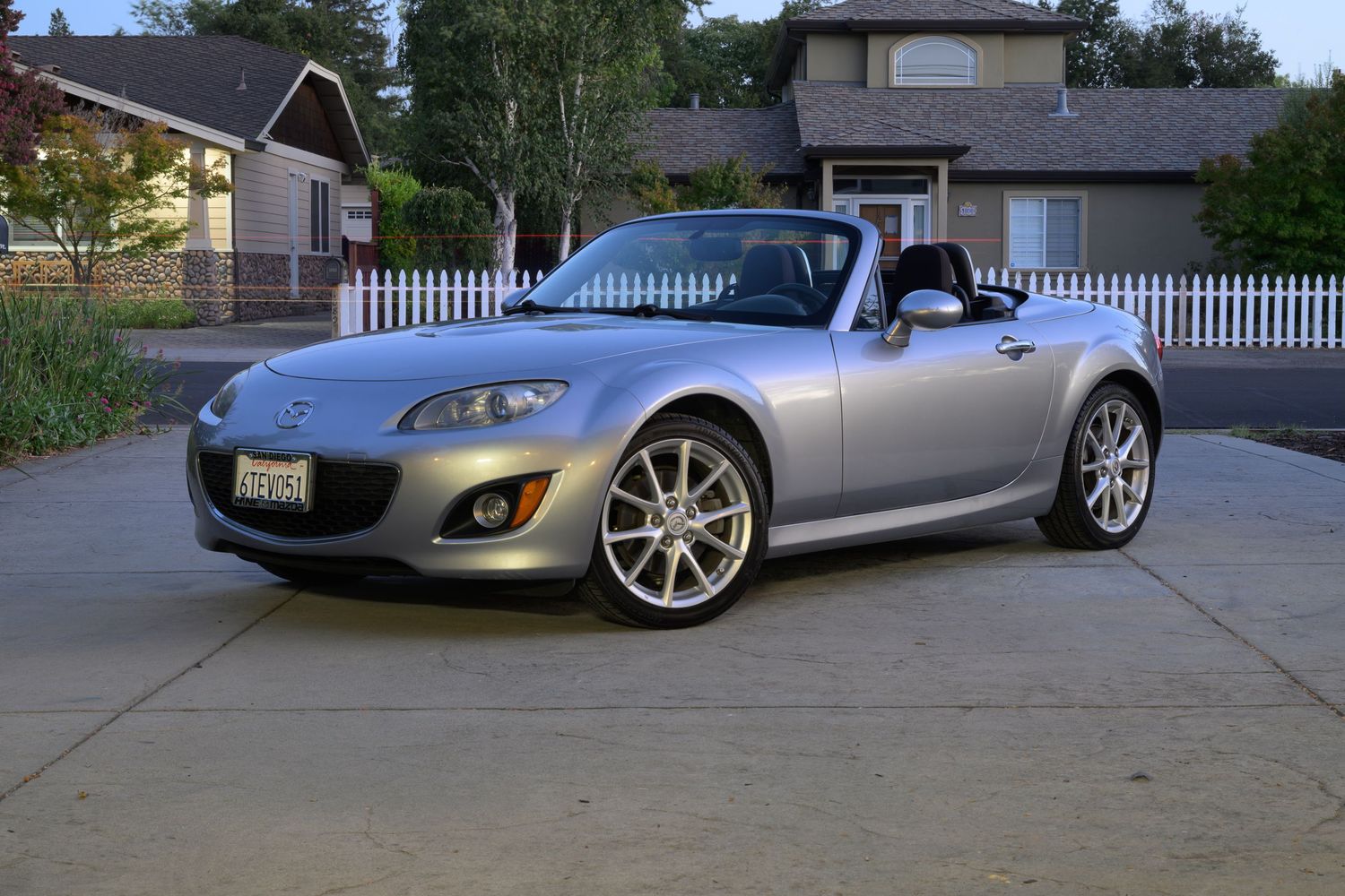 A silver Mazda Miata MX-5 sports car photographed in the evening with natural light on a suburban driveway.