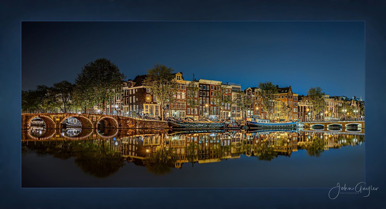 Illuminated canal houses and boats reflect in calm waters during twilight along Amsterdam's historic waterfront.