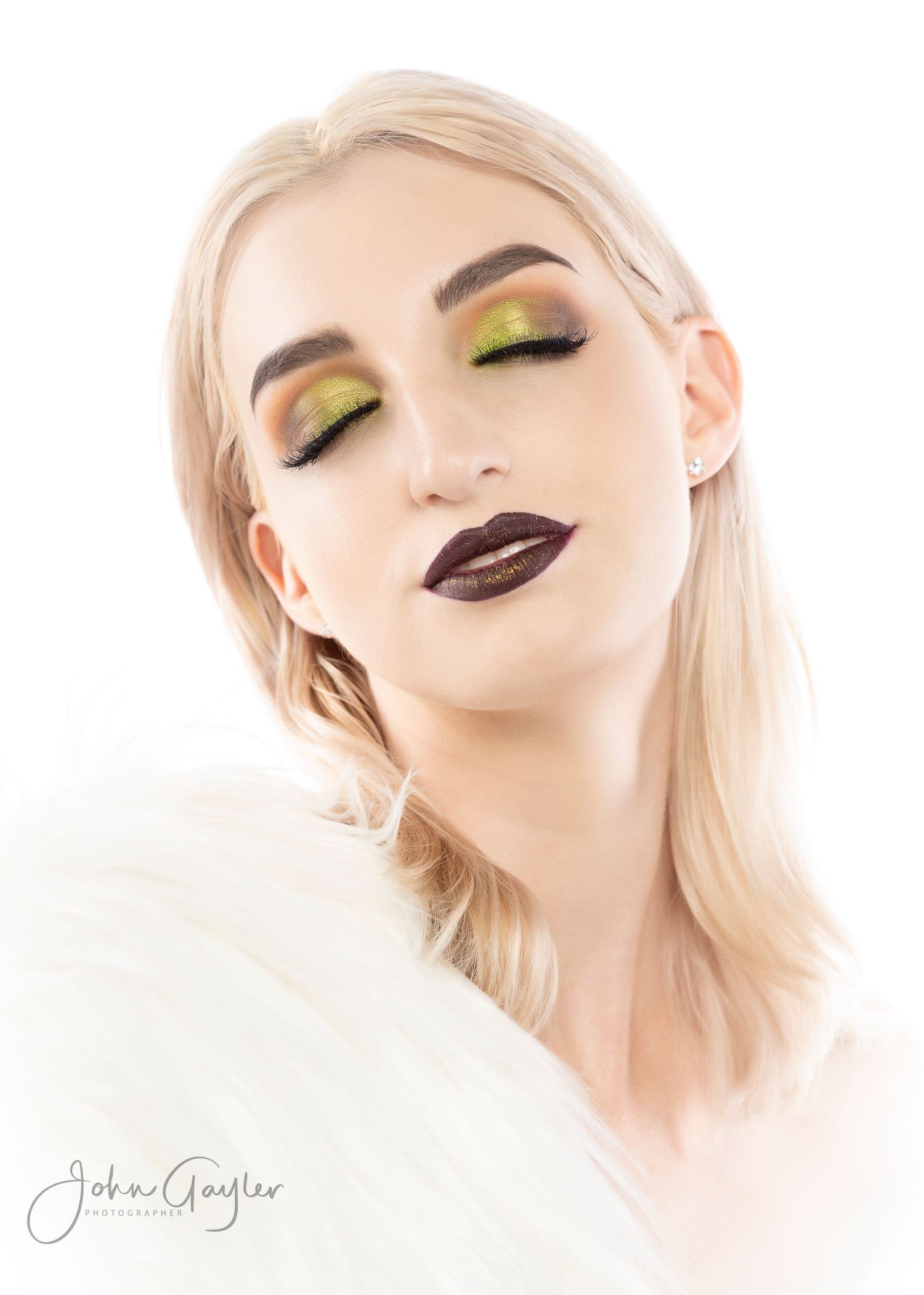 Beauty portrait featuring dramatic dark plum lipstick, yellow eyeshadow and platinum blonde hair against white background.
