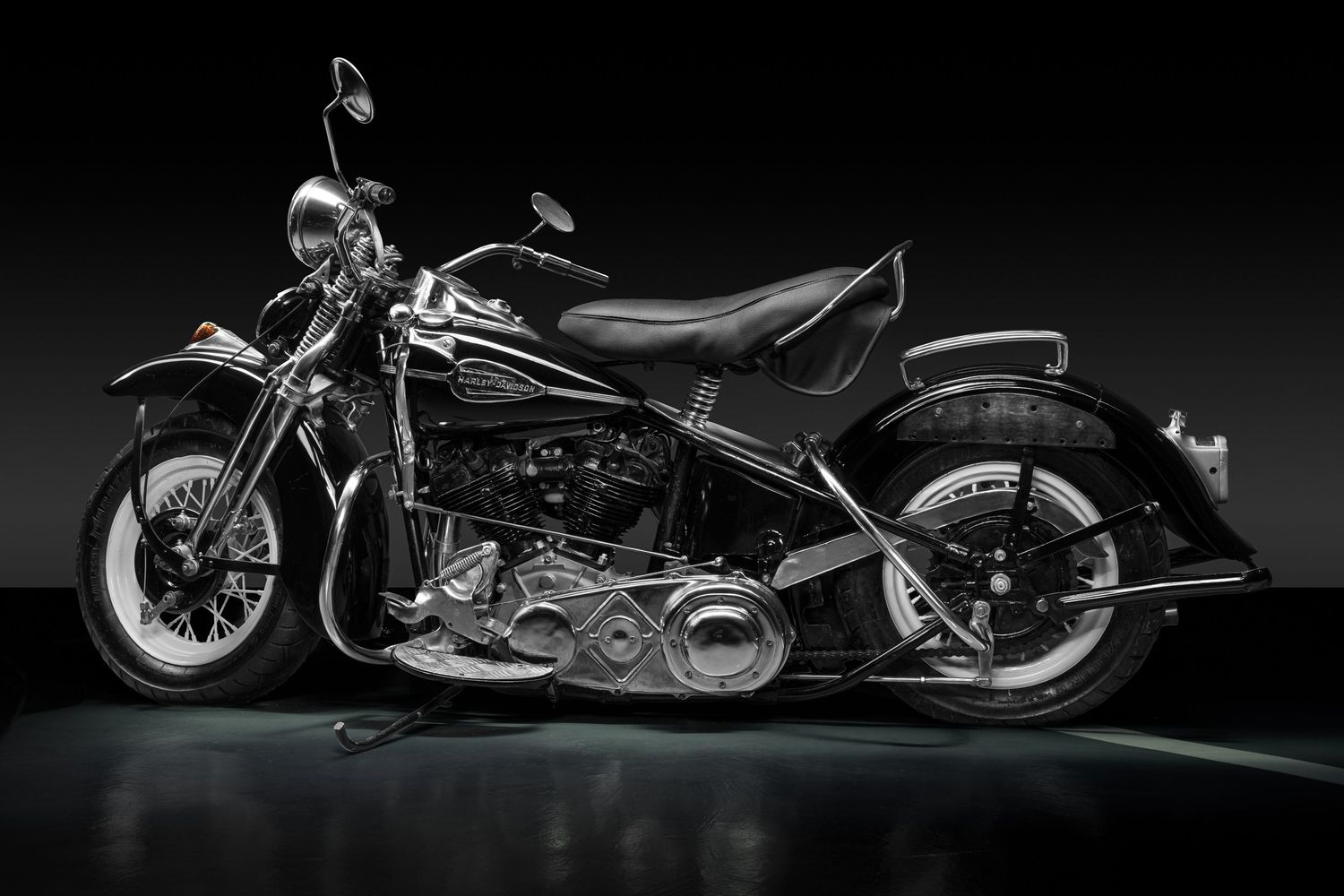 A sleek black 1940s classic Harley Davidson motorcycle light painted in a dark studio with dramatic lighting and reflective surfaces.