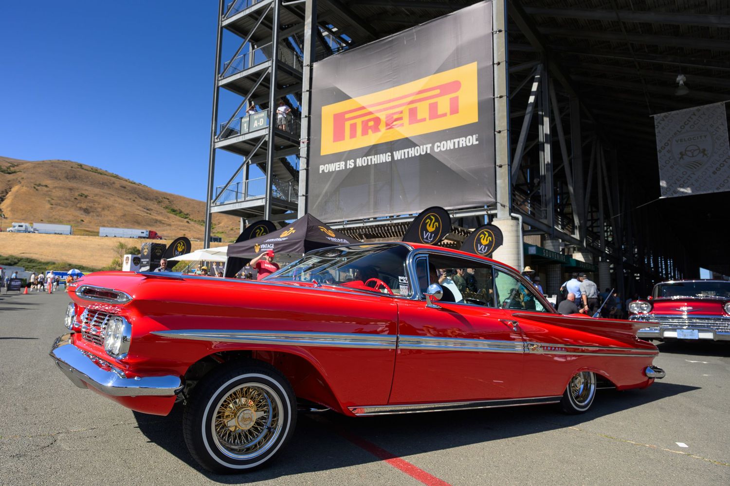 A vintage red convertible car with chrome trim and whitewall tires displayed at an outdoor automotive event held at the Sonoma Raceway.