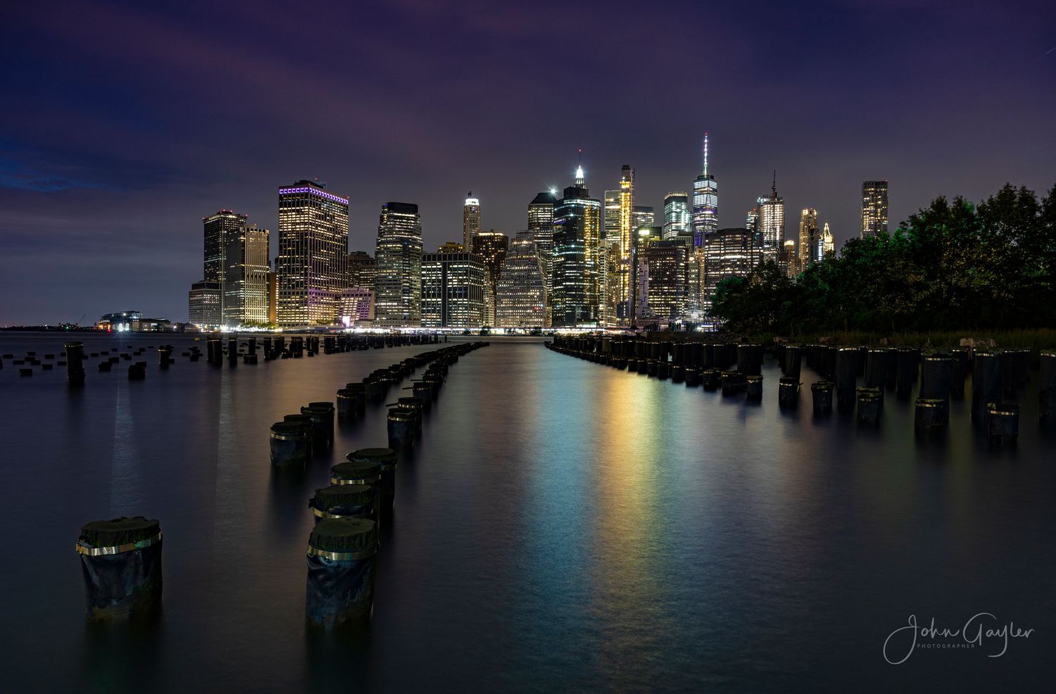 Nighttime cityscape of the New York skyline reflecting on the Hudson River with illuminated skyscrapers and World Trade Center Tower.