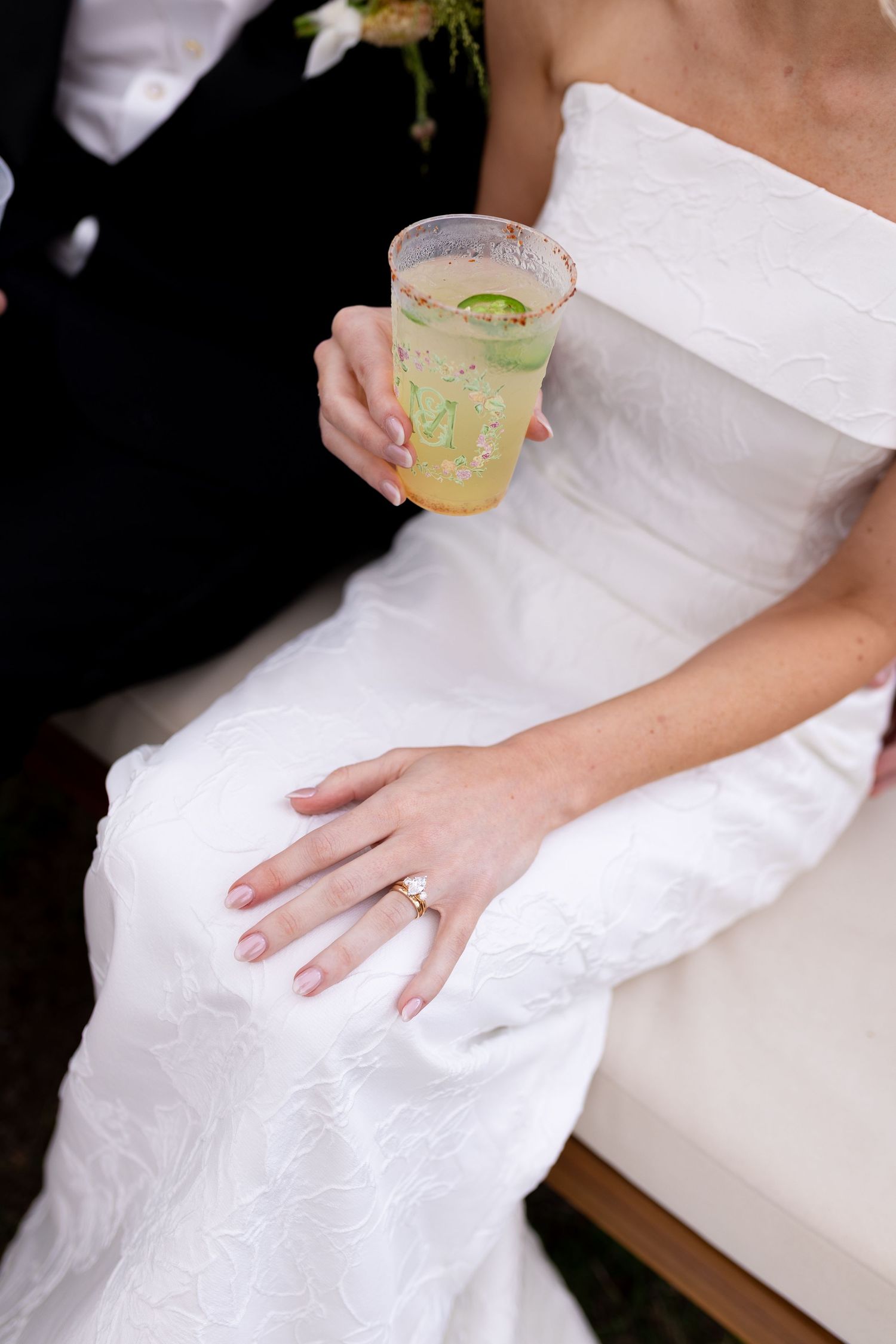 Close-up of bride's hand with ring holding citrus cocktail in white dress.