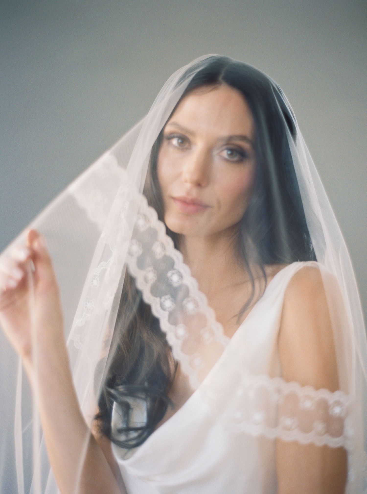 Bride wearing a delicate lace-trimmed tulle veil, holding it softly while posing in a white bridal gown.